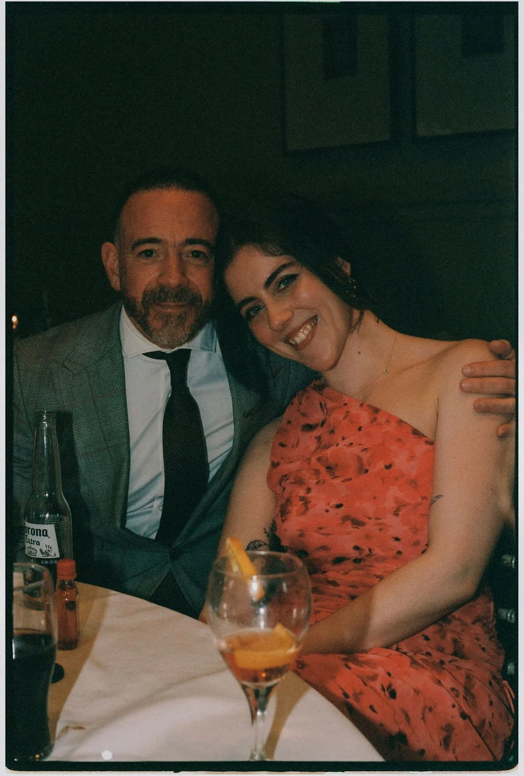 A man and woman sitting close together at a table, smiling. The man is wearing a checked suit and tie, and the woman is wearing an off-the-shoulder orange dress with black patterns. There are drinks and condiments on the table.