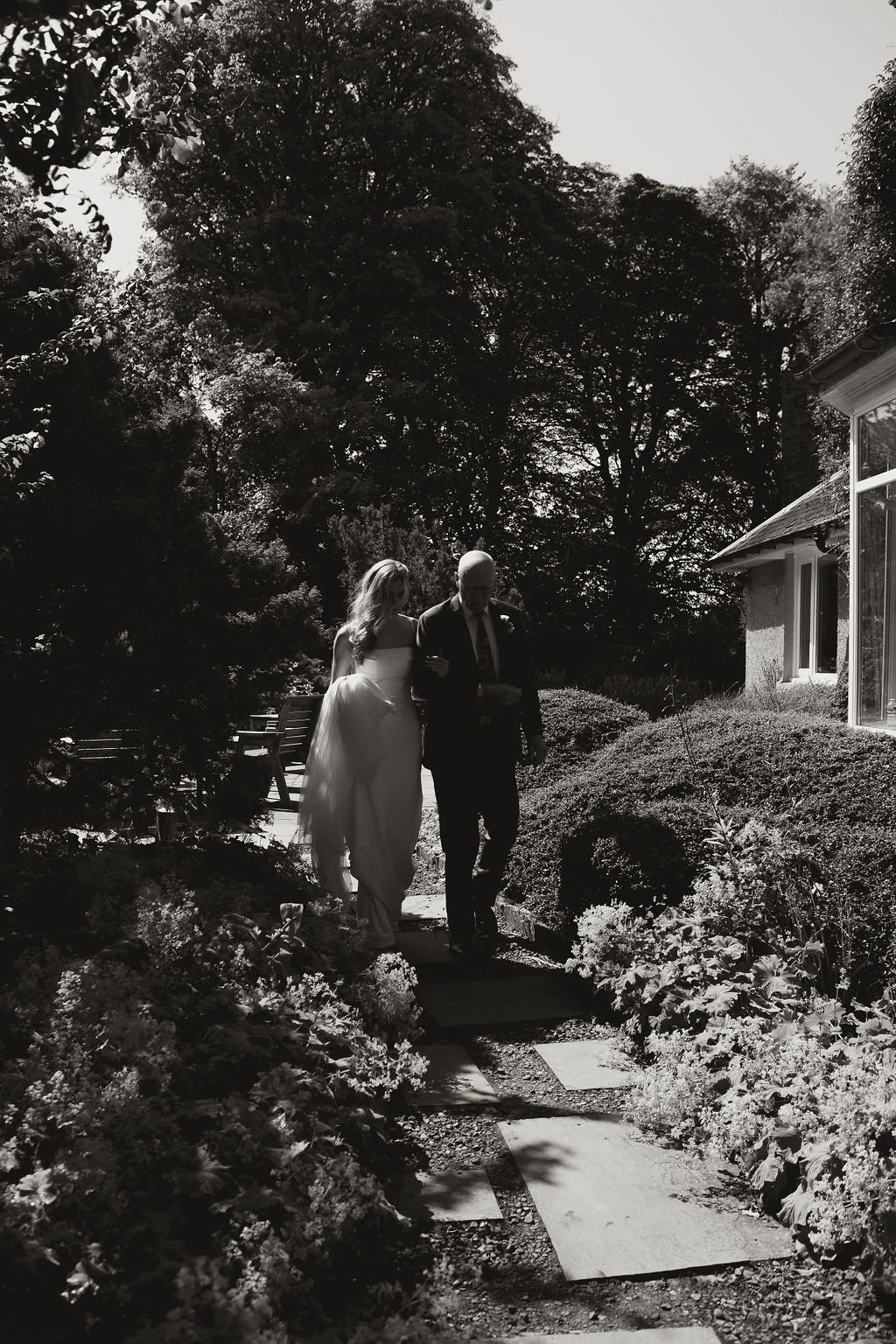 A bride and an elderly man walking arm-in-arm along a garden path outside a house with large windows. The scene is in black and white, with trees and bushes surrounding the area.
