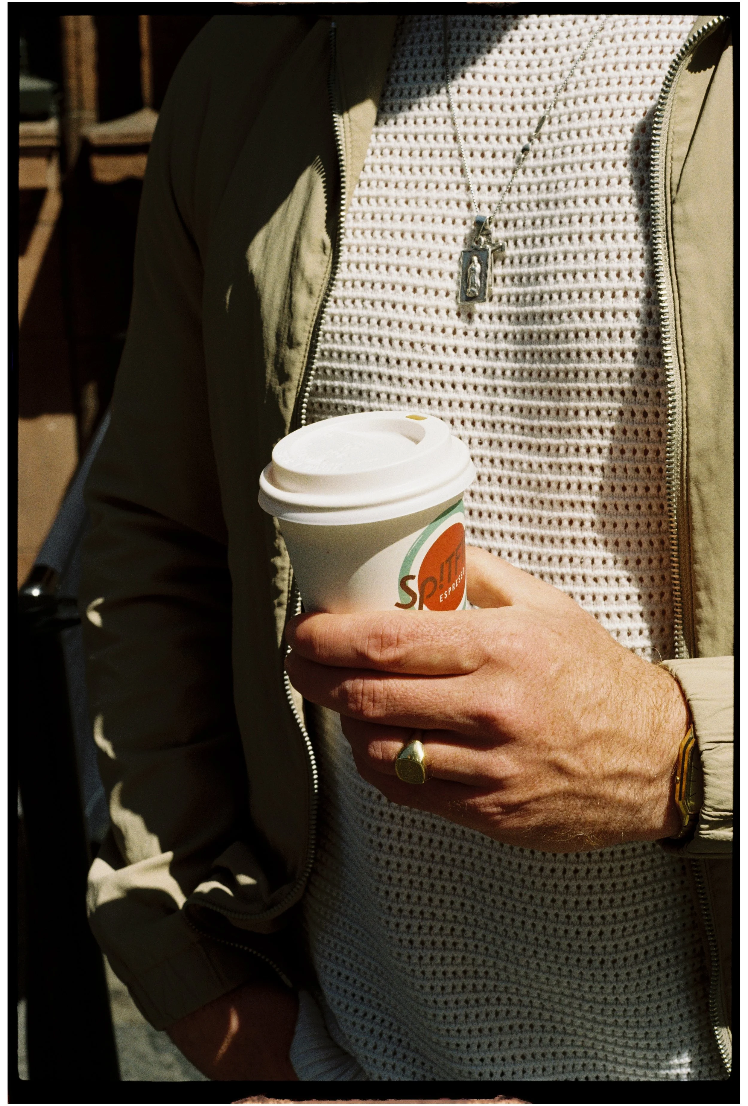 Person wearing a beige jacket and a white textured sweater, holding a disposable coffee cup, with a necklace and a gold wedding ring visible, on 35mm film.
