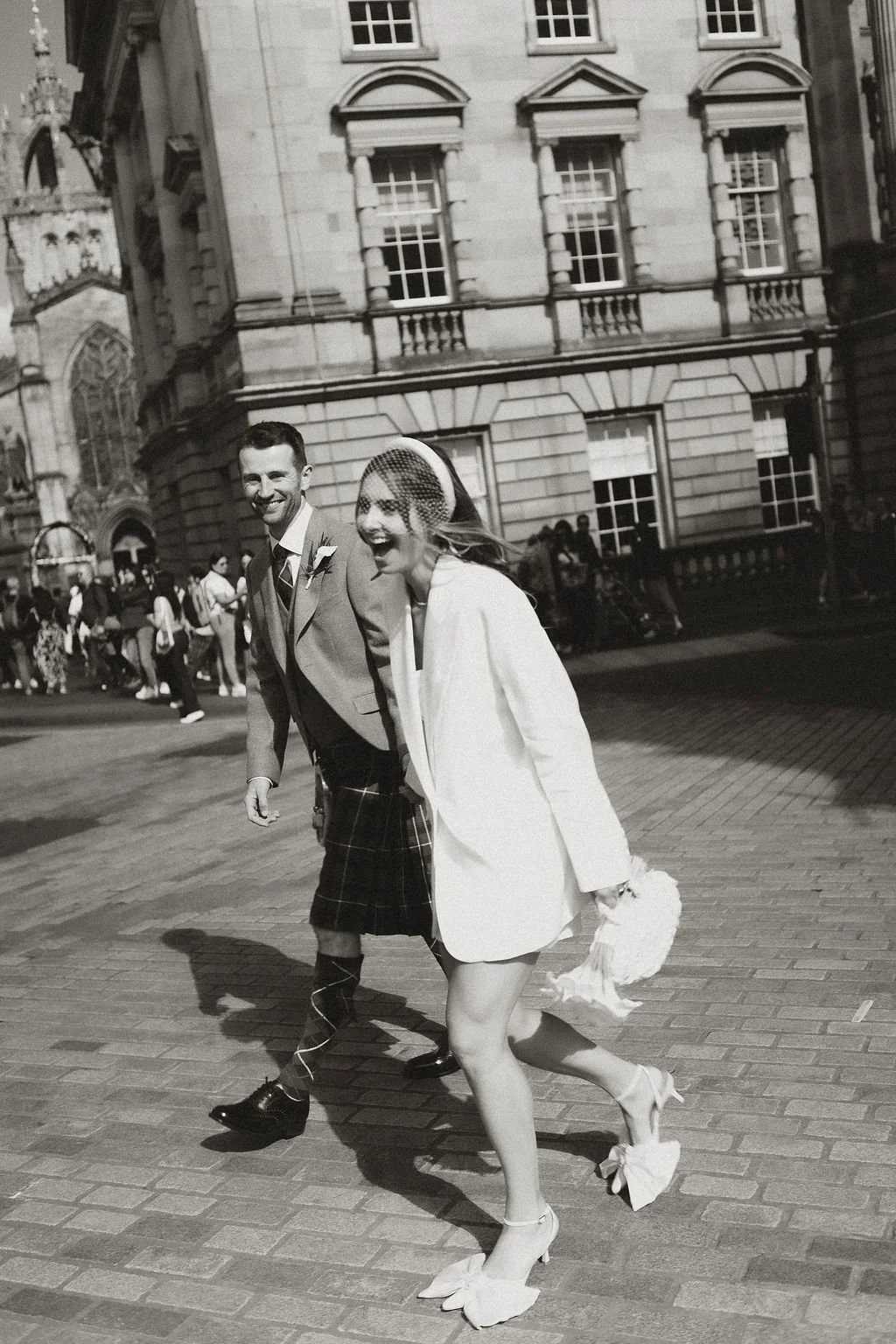 A bride and groom walking outside in an urban setting, smiling and laughing, with a crowd of people in the background. The bride is wearing a white coat, veil, and high heels, while the groom is in a suit and a kilt.
