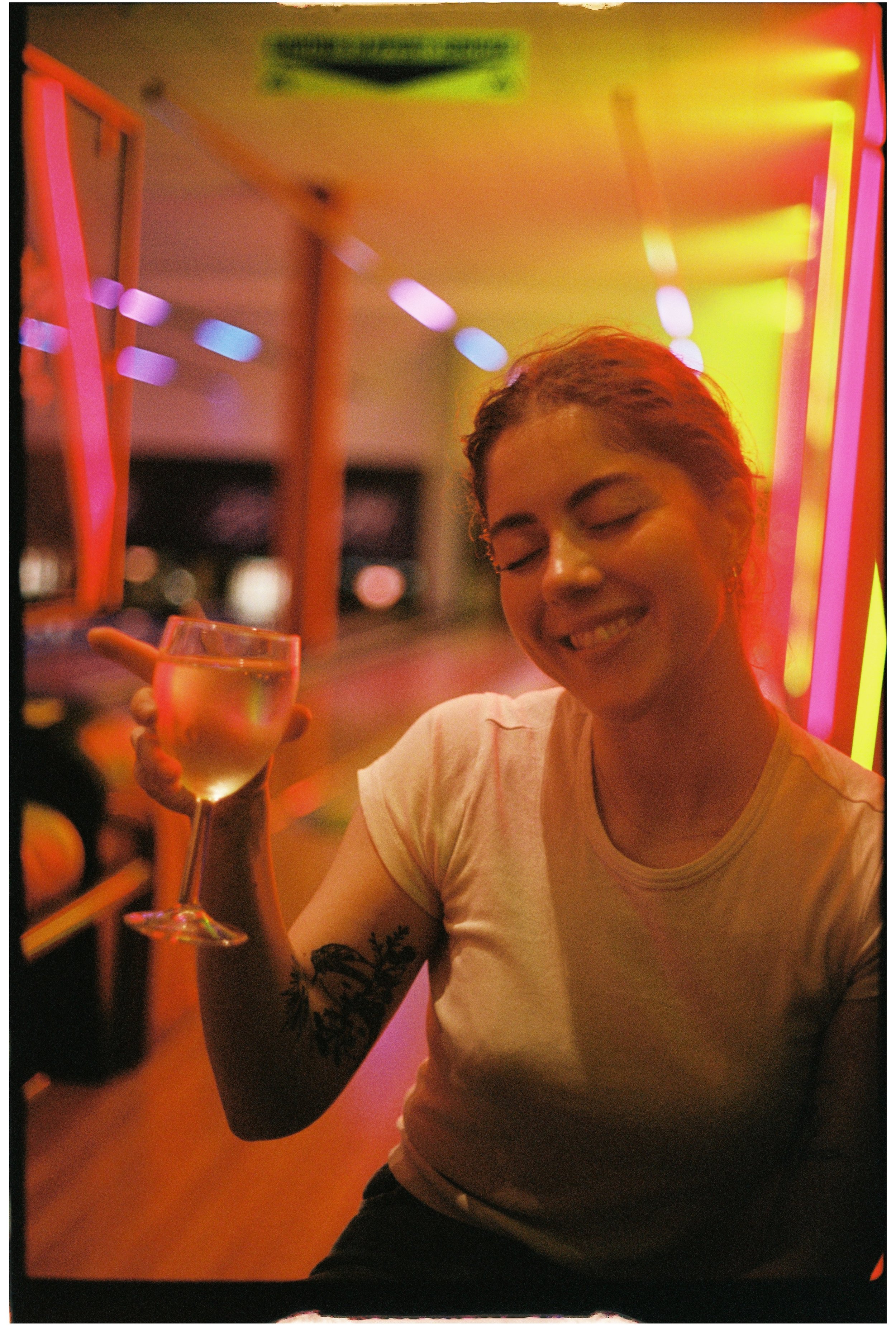 A woman with a tattoo on her arm holding a wine glass. She has her eyes closed and a big smile. The background features colorful neon lights in yellow, pink, and blue. Photographed on 35mm film.
