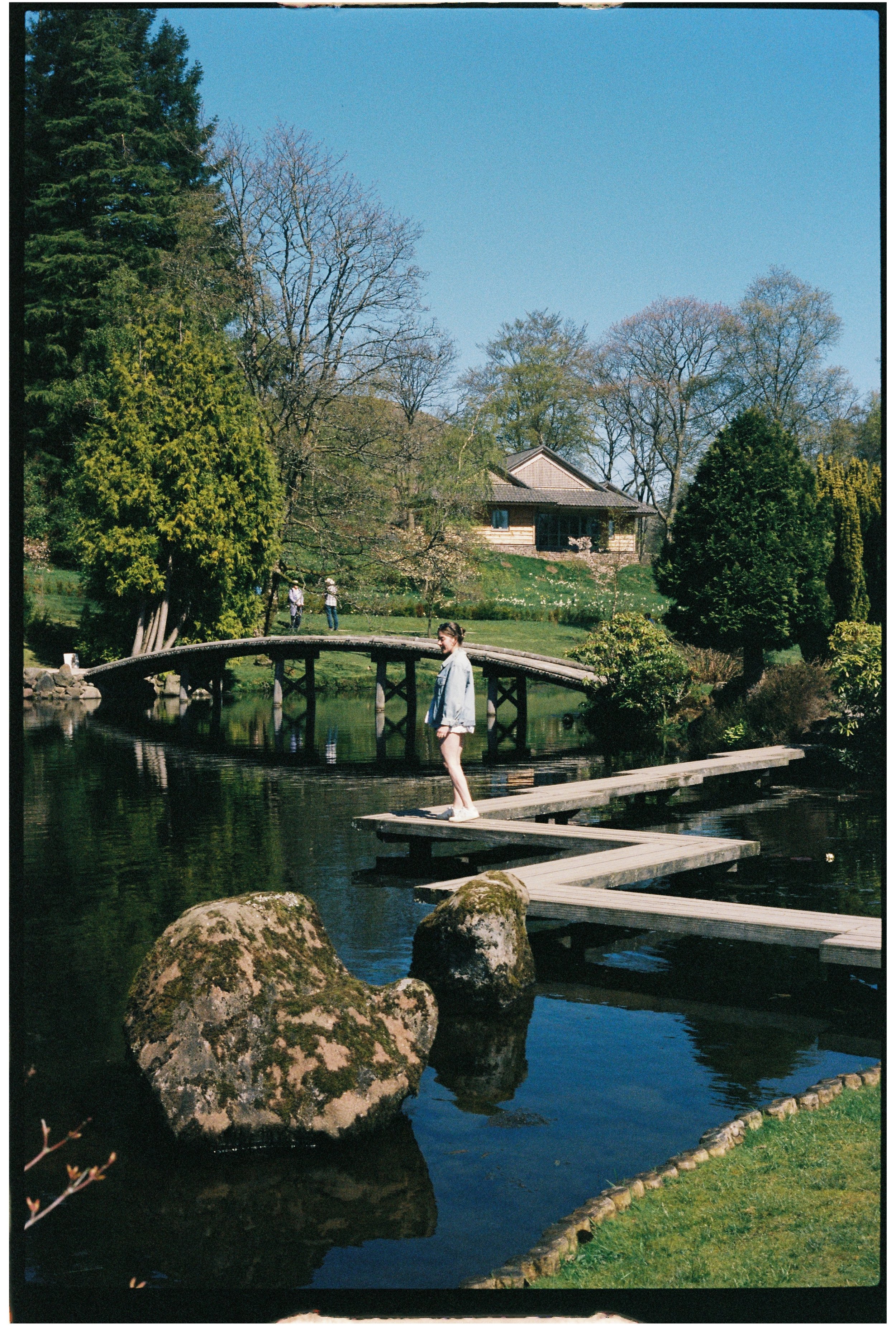 A person standing on a wooden dock over a small pond, with large rocks in the water and a lush garden with trees and a house in the background, on 35mm film.