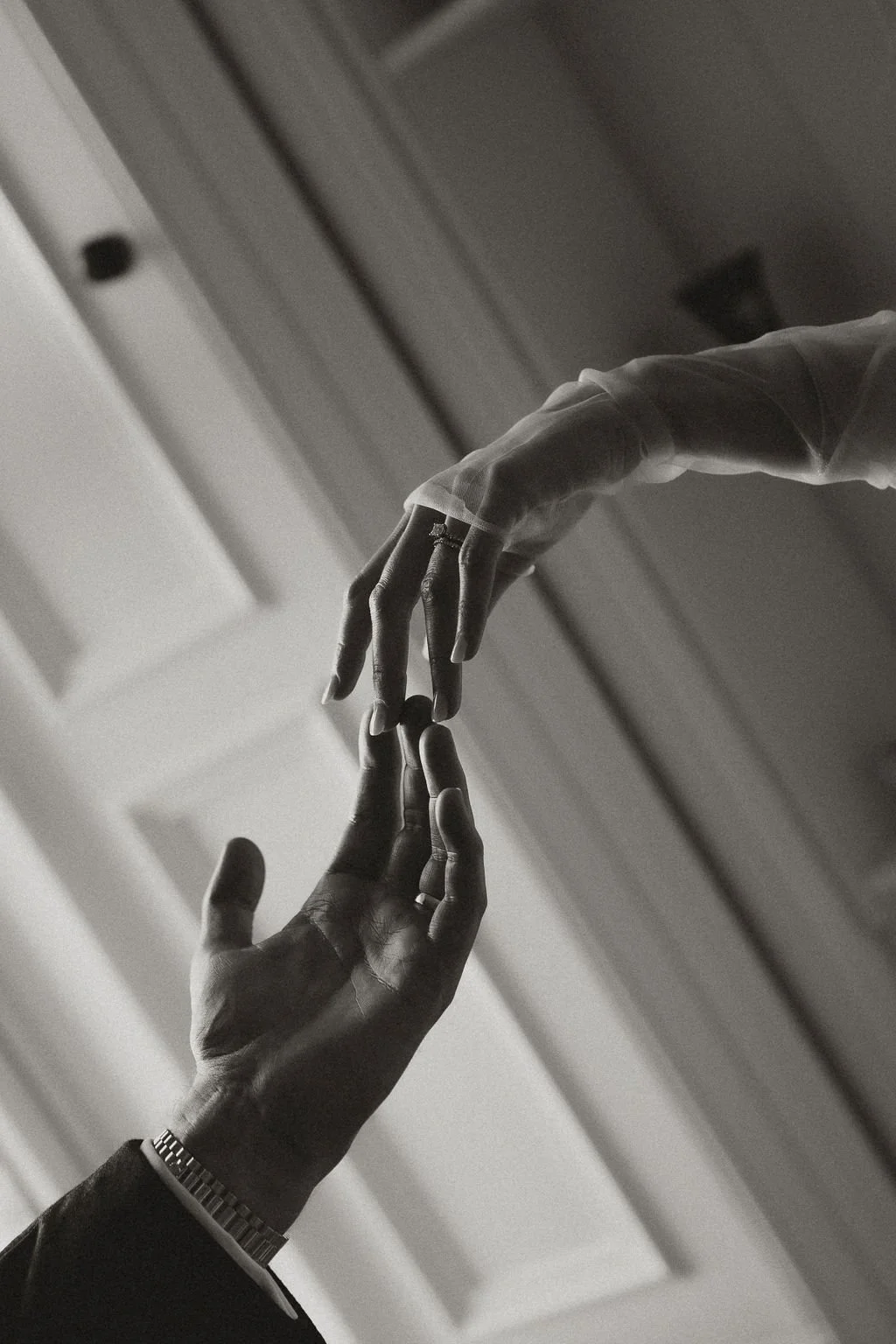 Black and white photo of two hands reaching towards each other, with fingers almost touching, against a background of a door frame and ceiling.