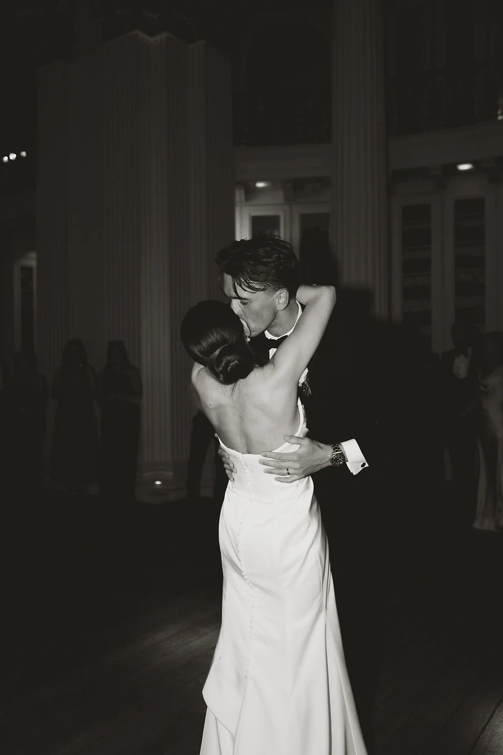 A black and white photo of a bride and groom sharing a dance at their wedding reception, with the groom in a tuxedo and the bride in a strapless gown, embracing and kissing.