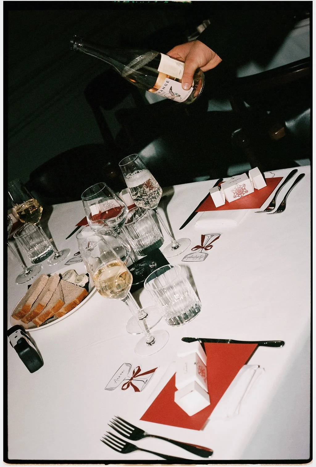 A table set for a celebration with wine glasses, a plate of sliced bread, utensils, small gift boxes, and holiday-themed decorations on red placemats.