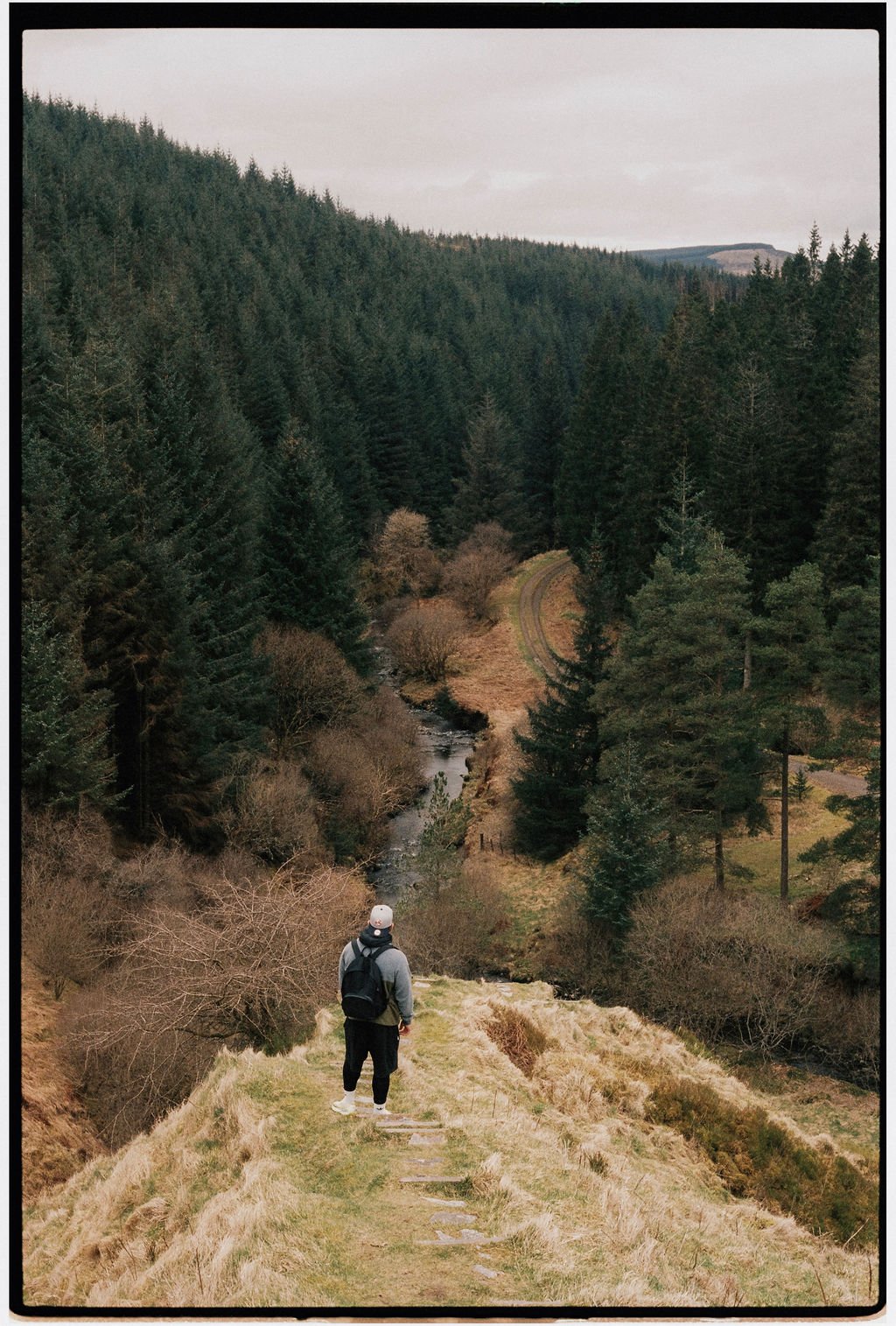 A person with a backpack standing on a grassy trail overlooking a forested valley and a small stream, with a mountain range in the background.