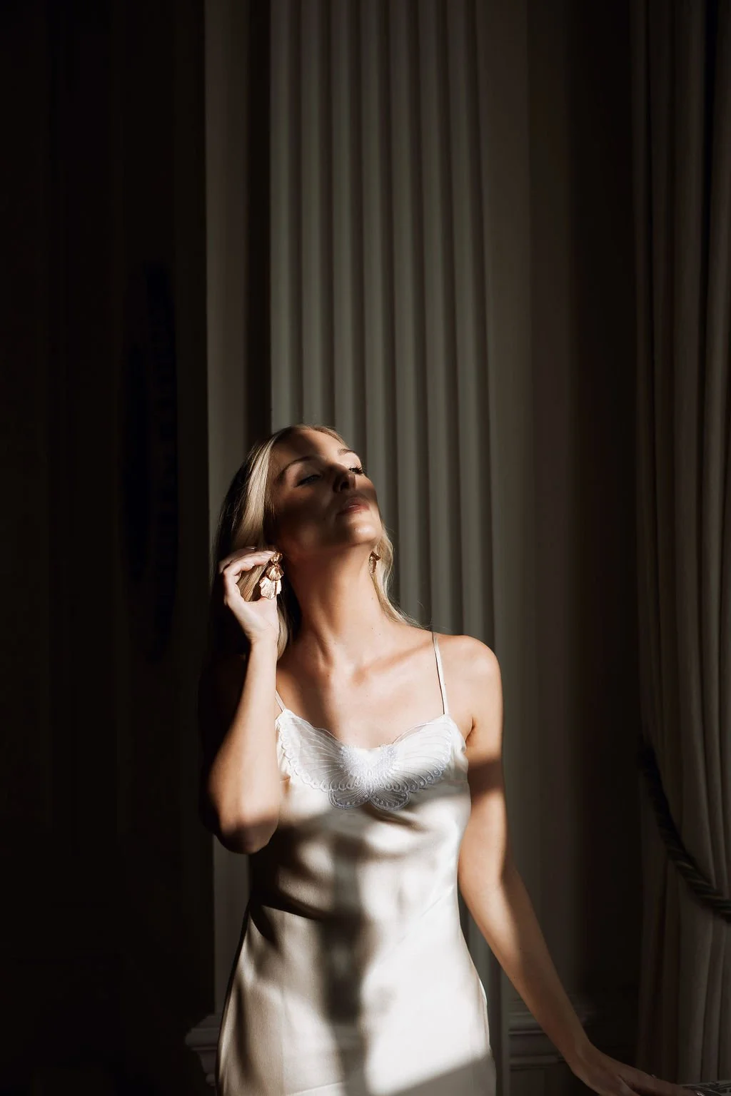 A woman is standing next to a window, adjusting a large earring. She is wearing a silky, white dress with spaghetti straps and a butterfly detail on the chest. Sunlight streams in, casting shadows on her face and the background curtains.