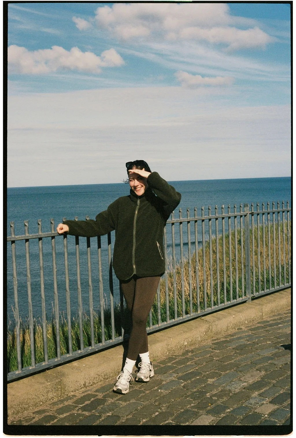A woman standing by a metal railing on a coastal walkway, smiling and shielding her eyes from the sun, with the ocean and partly cloudy sky in the background.