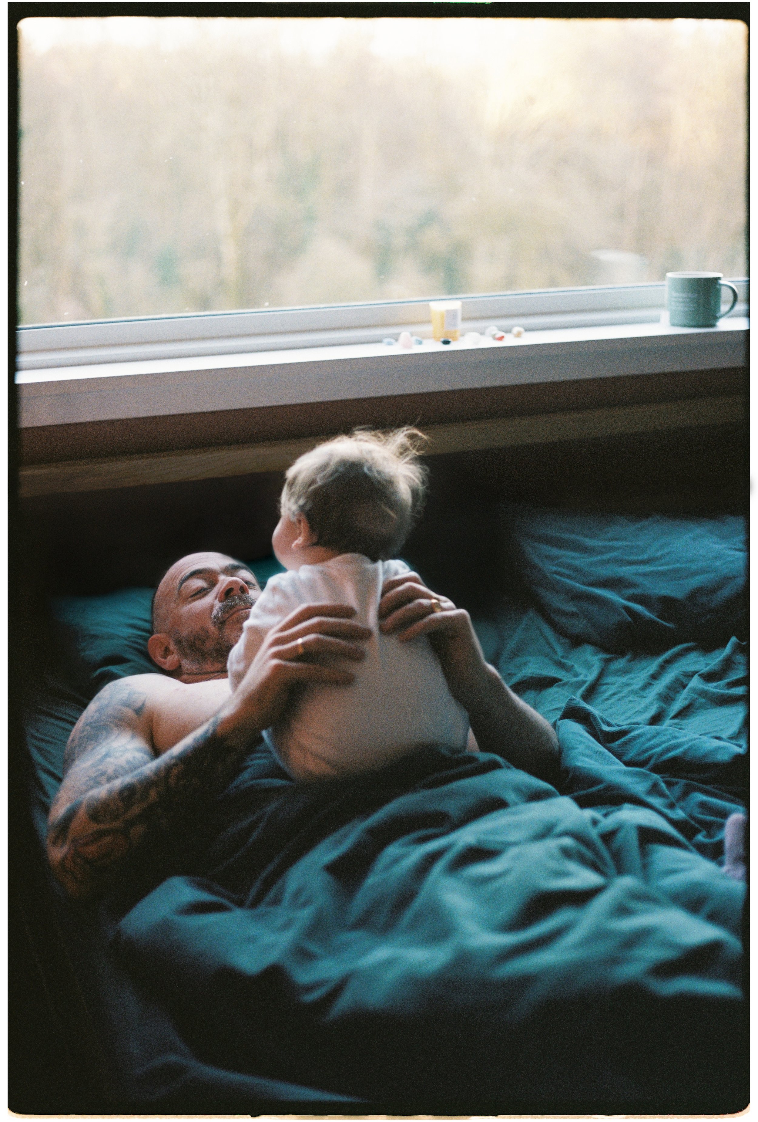 A man with tattoos lying in bed, holding a young child who is facing him. The man is looking at the child with his eyes closed, and the child is looking at the man. The bed has dark blue sheets, and there is a window in the background with a window s