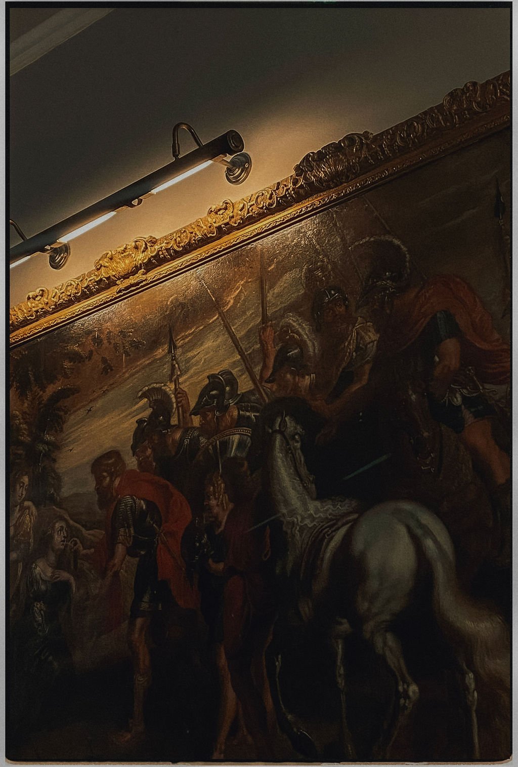 A historical painting depicting a battle scene with soldiers on foot and on horses, carrying weapons like spears and swords, with dark, somber tones and ornate gold details on the upper margin.