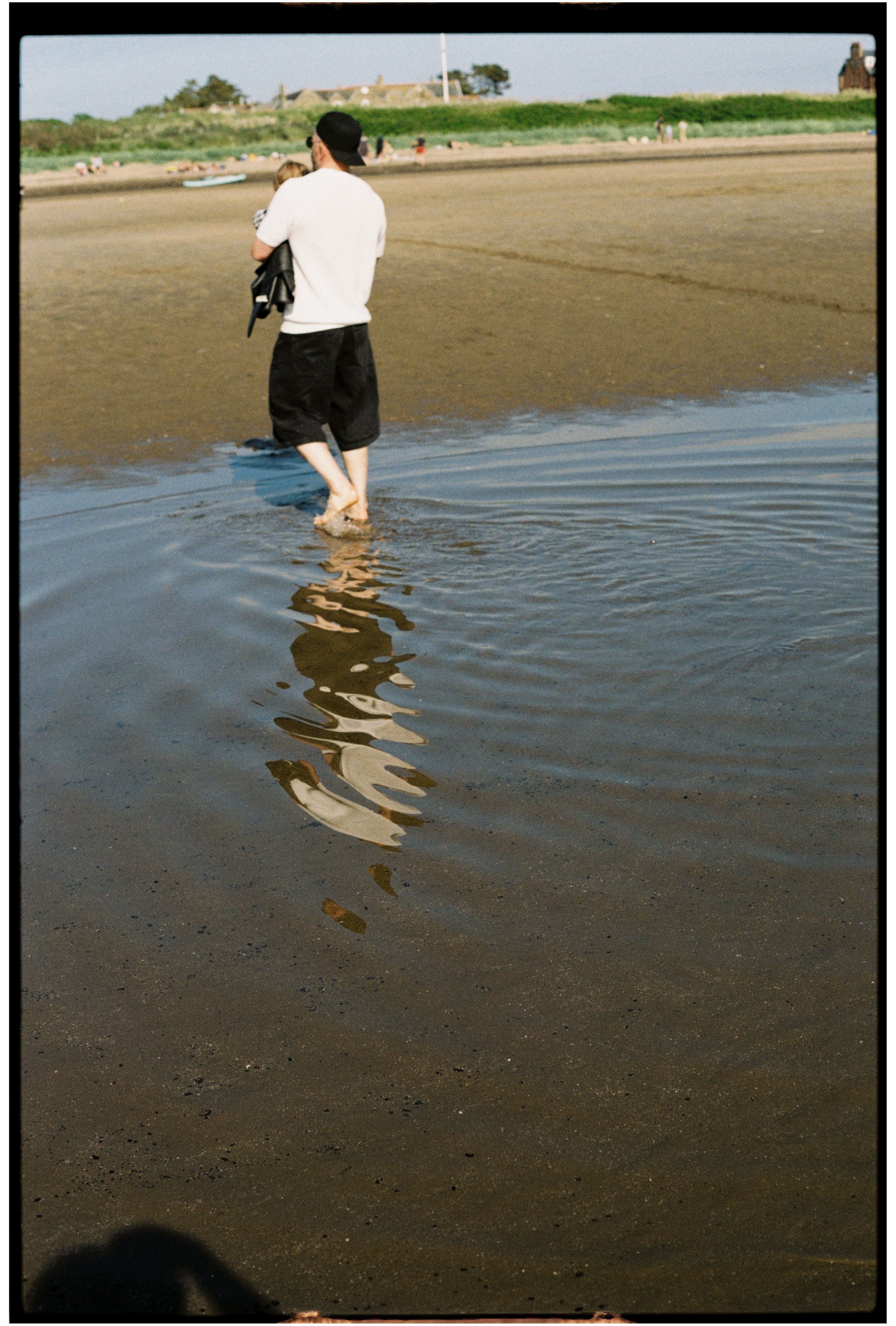 A person walking on a beach with shallow water, holding a child in their arms. The reflection of the person and the child can be seen in the water. In the background, there is a green grassy area, some coastline buildings, and a few people in the dis