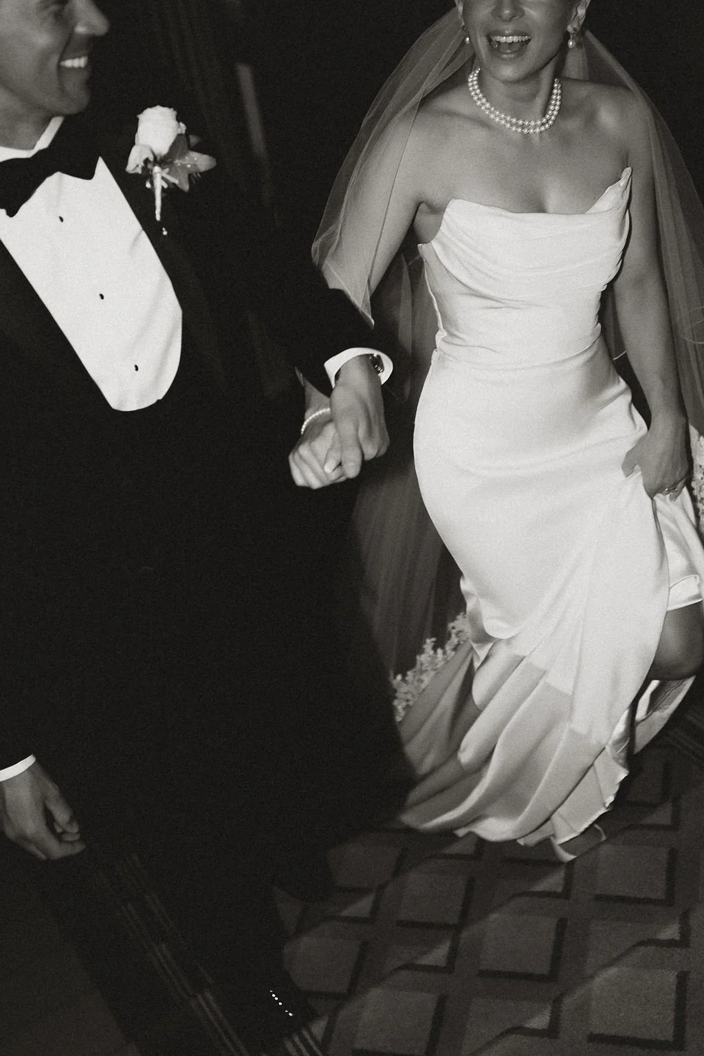 A bride and groom holding hands during their wedding dance, with the bride wearing a strapless wedding gown and pearl necklace, and the groom in a tuxedo with a boutonniere.
