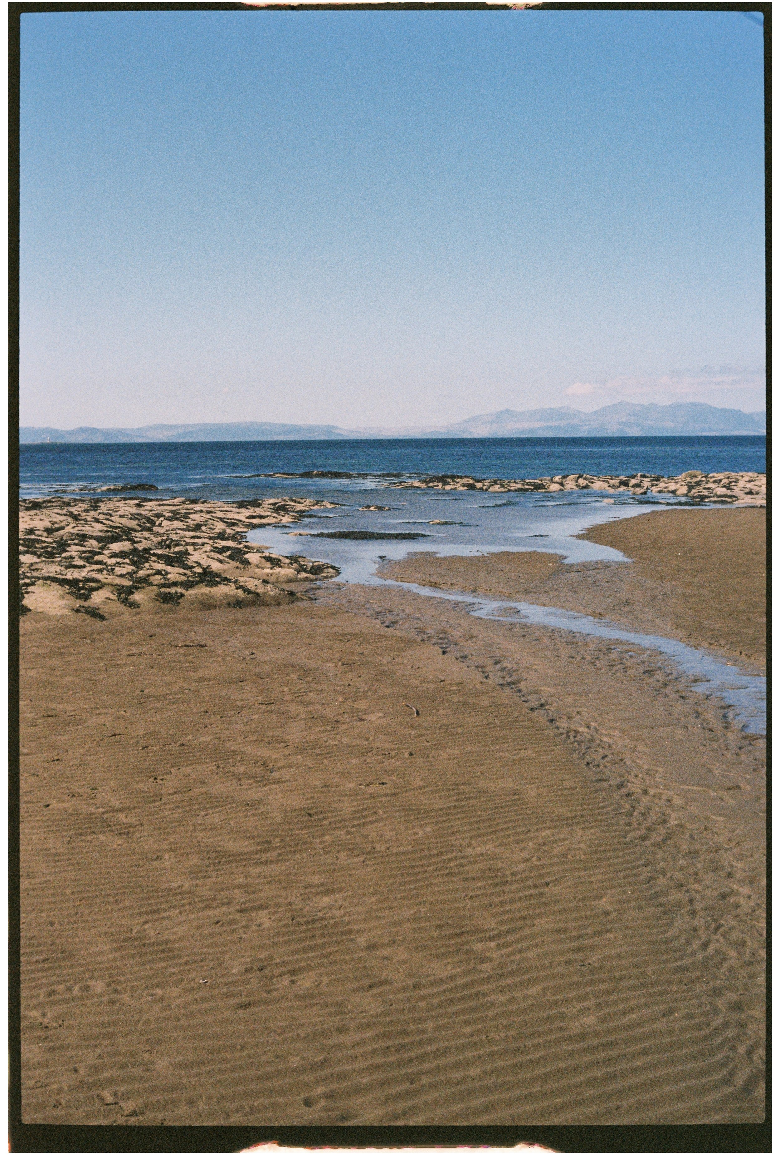A sandy beach with ripples in the sand, rocky shoreline extending into the ocean, calm water, and distant landforms under a clear blue sky. Photographed on 35mm film.