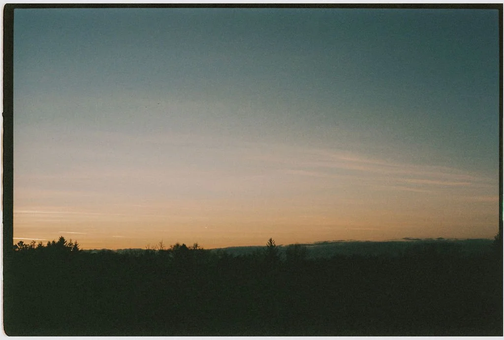 A landscape photo of a sunset or sunrise over a flat, rural area with trees on the horizon and a clear sky with pastel colors.