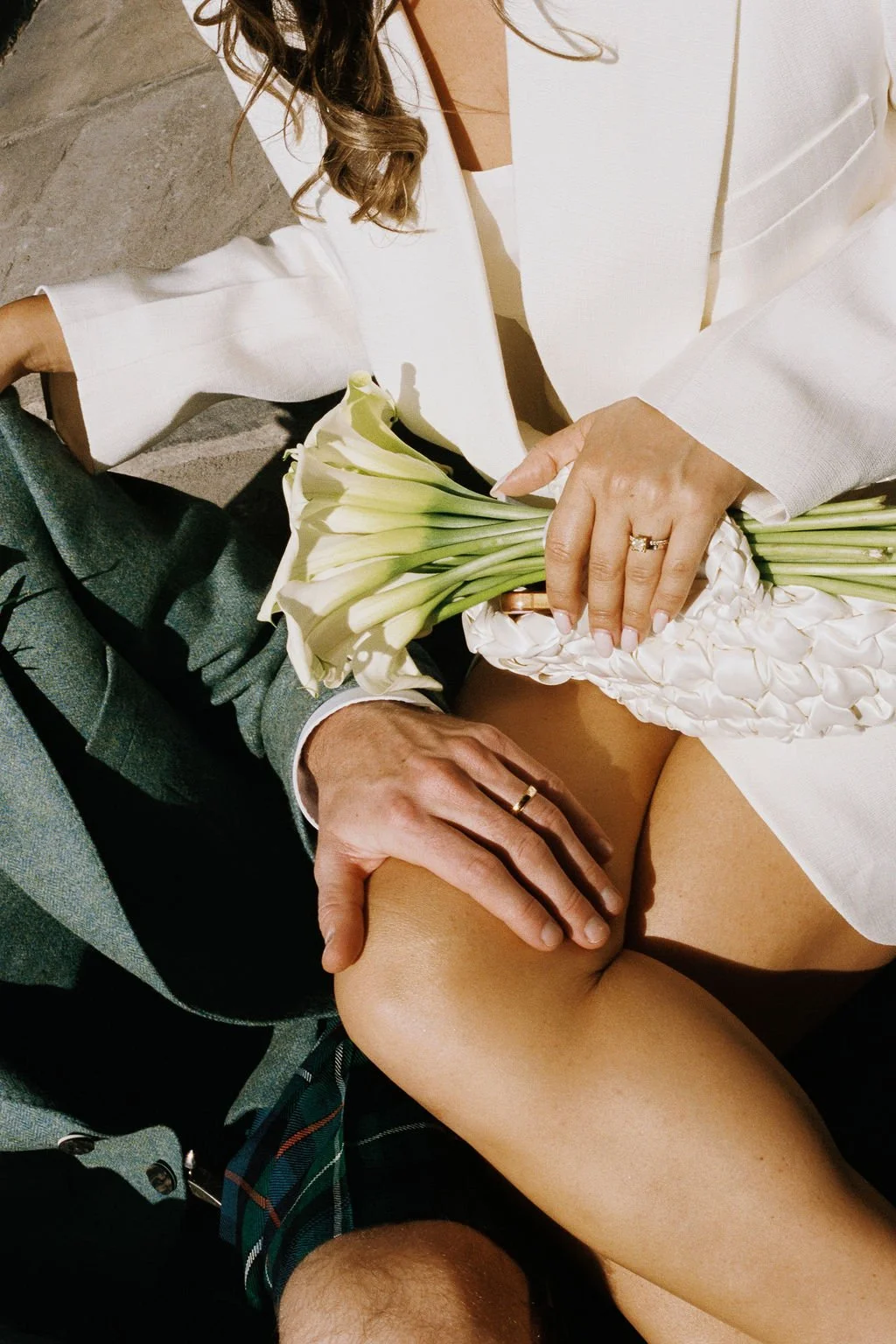A man in a gray suit with a plaid tie holding a bouquet of white calla lilies, with a woman in a white blazer and skirt sitting on his lap, placing her hand on his knee, both wearing rings.