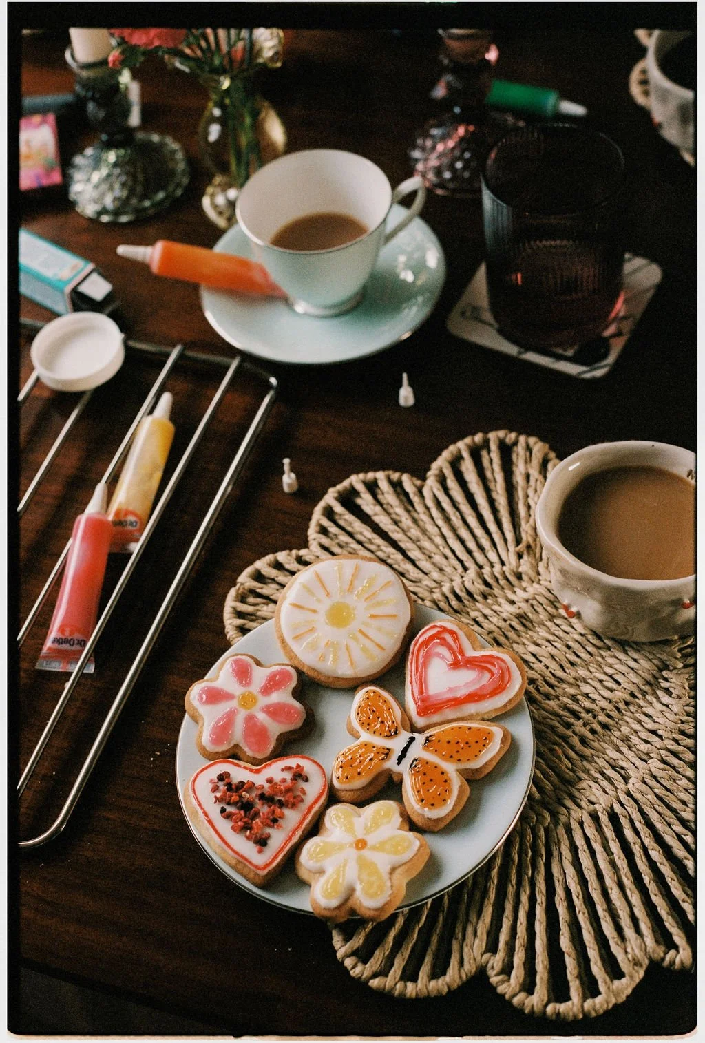 Assorted decorated cookies on a white plate, cups of coffee, a glass of dark beverage, and various items on a dark wooden table.