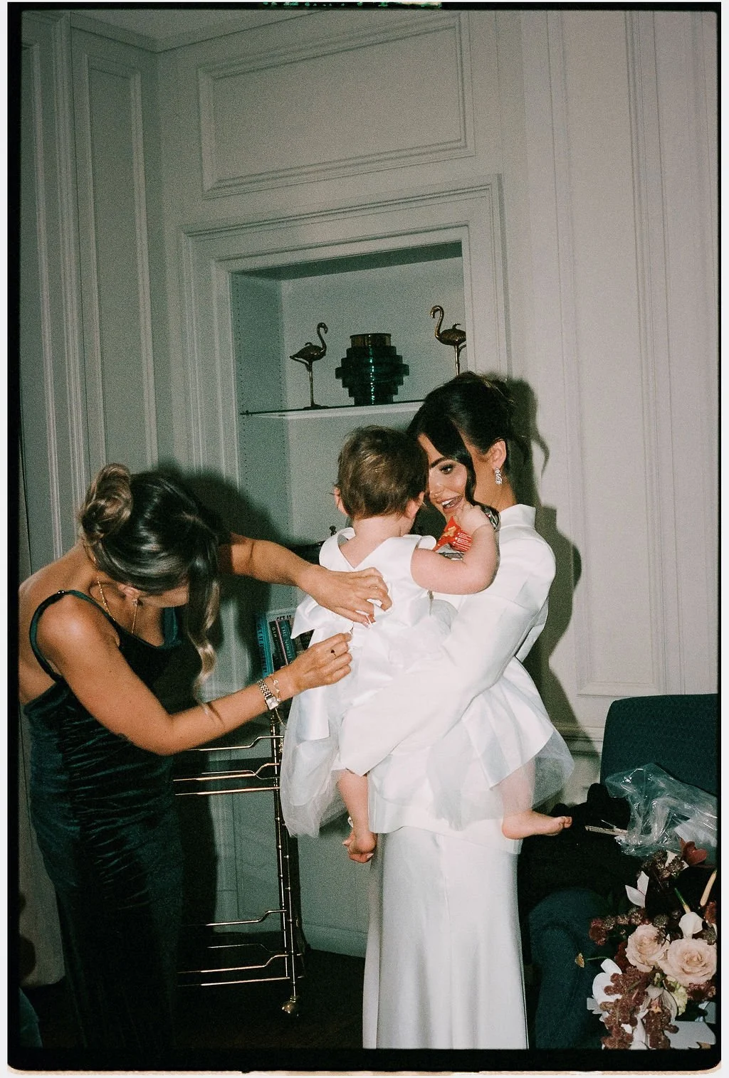 A woman in a white dress holding a young girl in a white dress, smiling at each other, while another woman adjusts the girl's dress in a room with white paneled walls and decorative items on shelves.