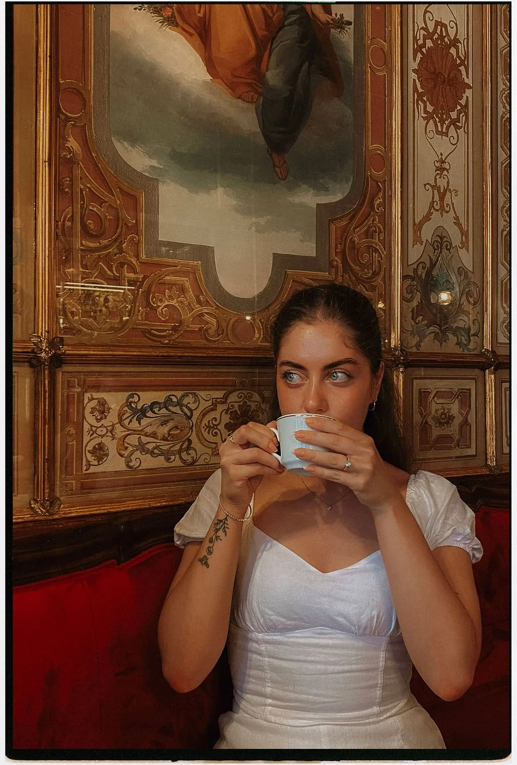 A young woman with dark hair and blue eyes drinks from a white cup in a richly decorated room with ornate wooden paneling and a large painting or fresco on the wall behind her.