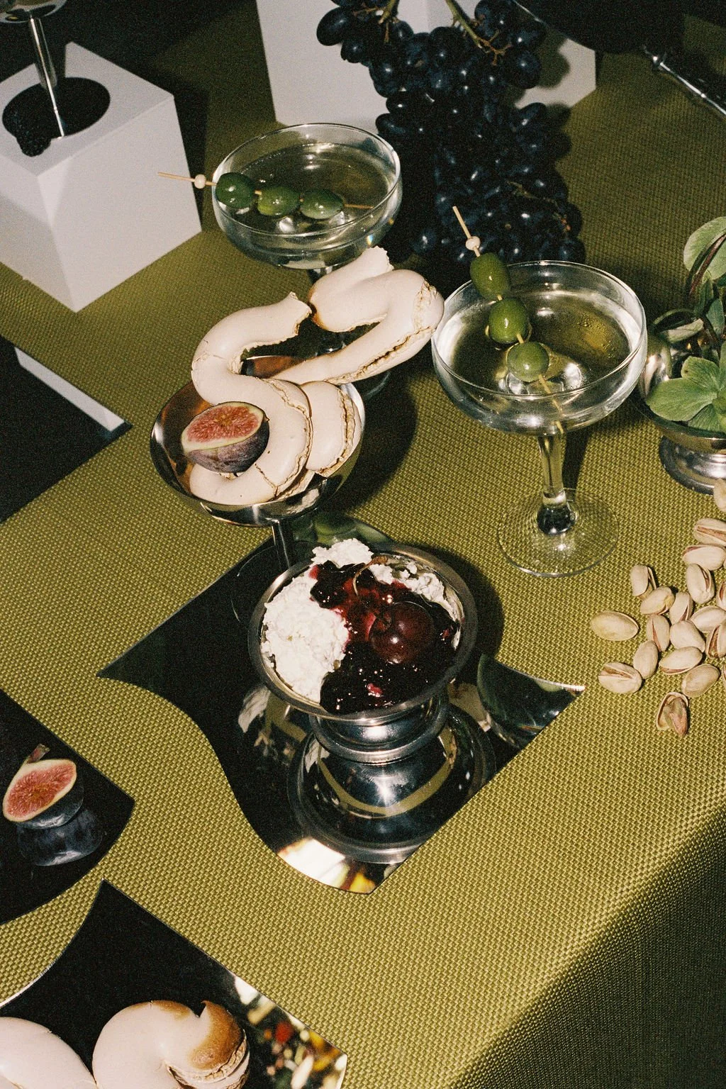An assortment of desserts and fruits on a table with a green tablecloth, including a bowl of whipped cream with cherries and berry compote, two cocktail glasses with green olives, and a plate of halved figs with pistachios and grapes in the backgroun