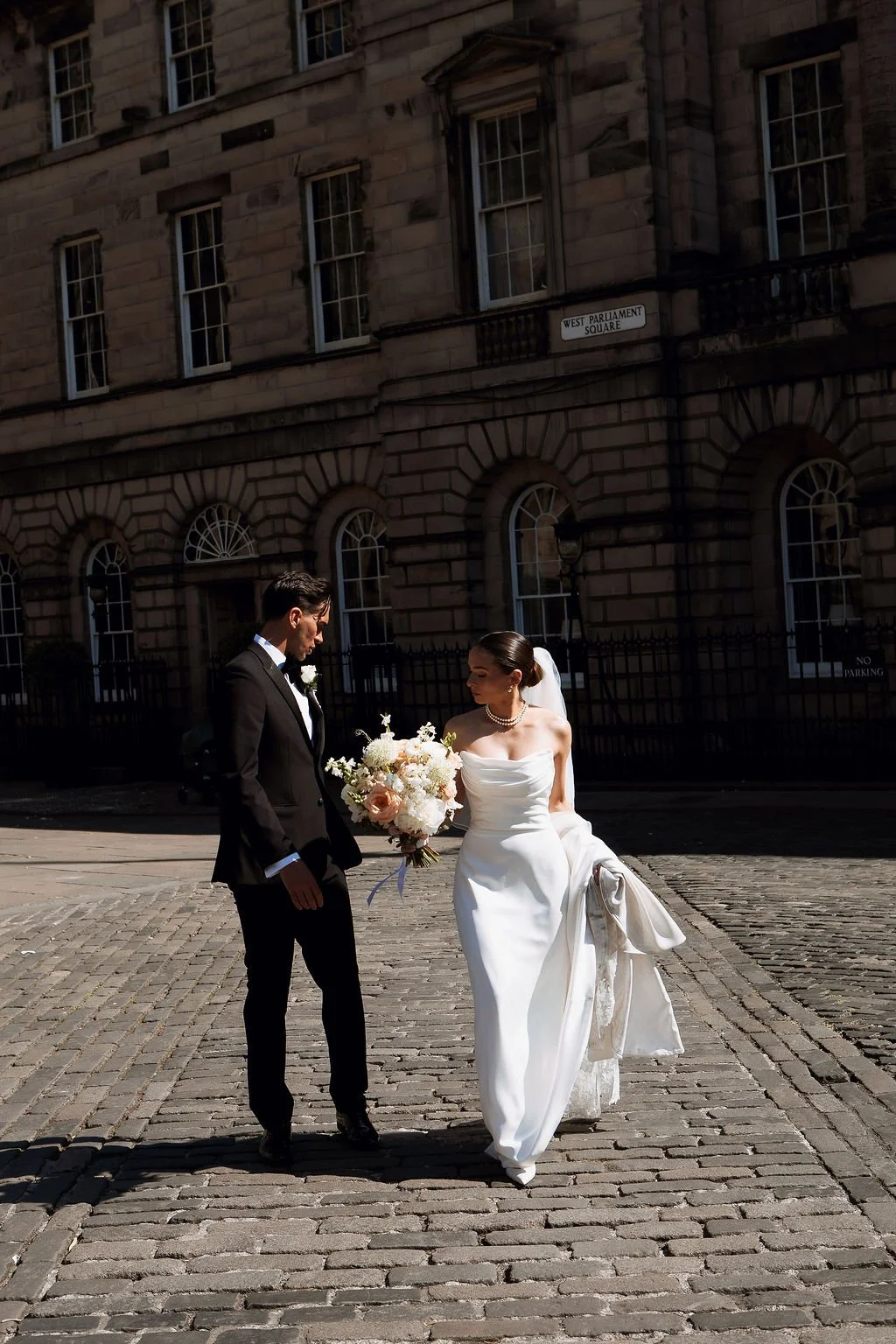 A bride in a white wedding dress holding a bouquet standing with a groom in a black tuxedo on a cobblestone street in front of a historic building.