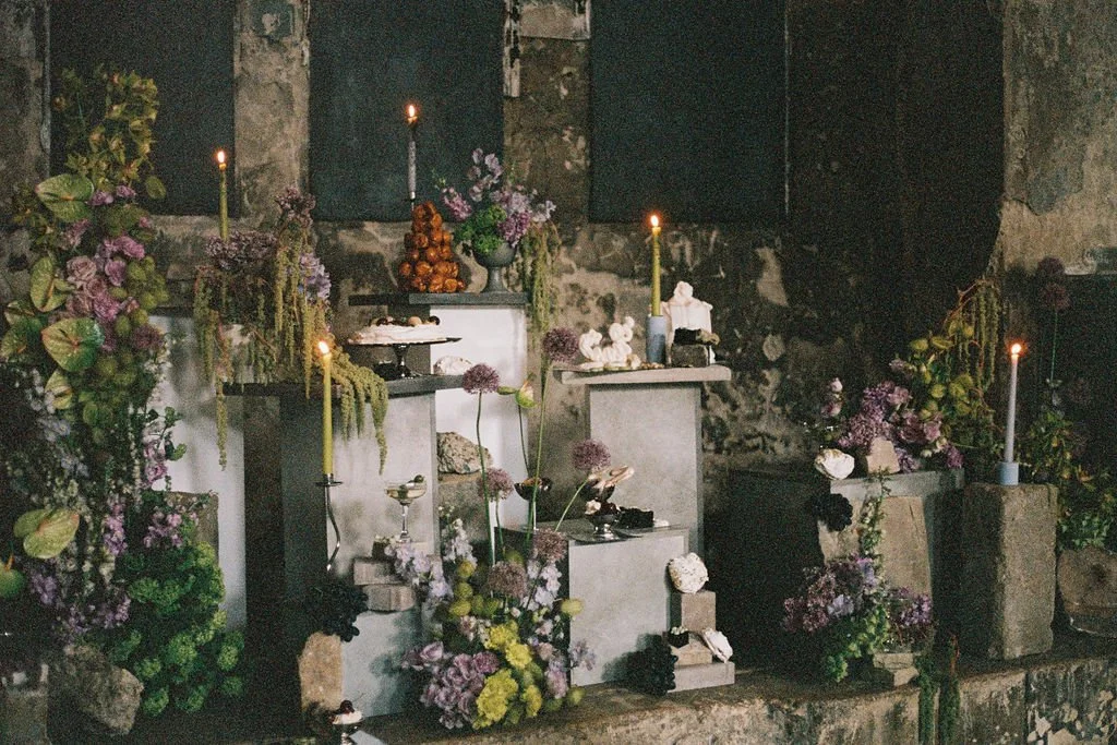 A small altar with lit yellow candles, surrounded by purple and green floral arrangements, set against a rustic stone wall.