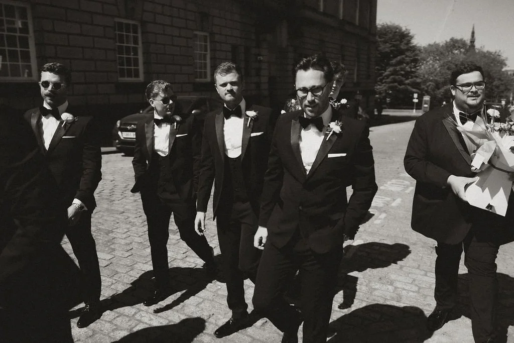 Five men in tuxedos walking on a cobblestone street, some wearing sunglasses, in black and white photo.