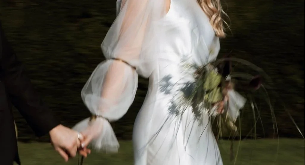 A bride in a white wedding dress holding a bouquet of flowers and a groom in a dark suit holding her hand during a wedding ceremony.