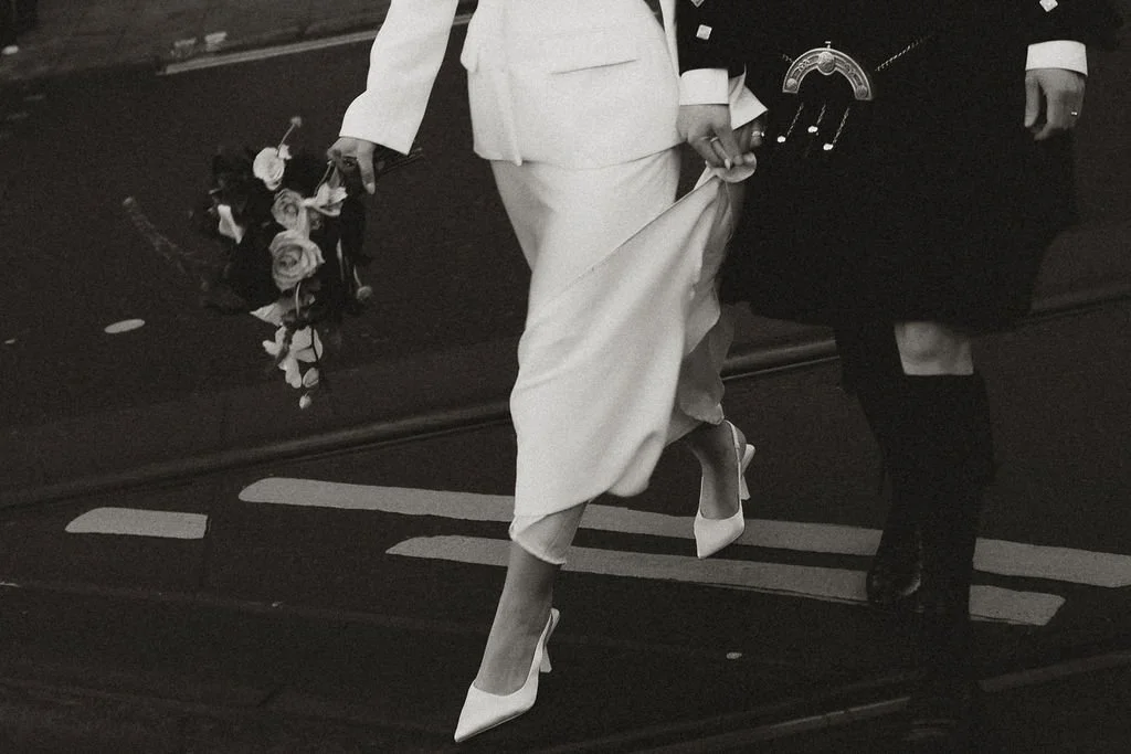 People walking on a city street, one carrying a bouquet of flowers, and wearing formal attire, including high heels.