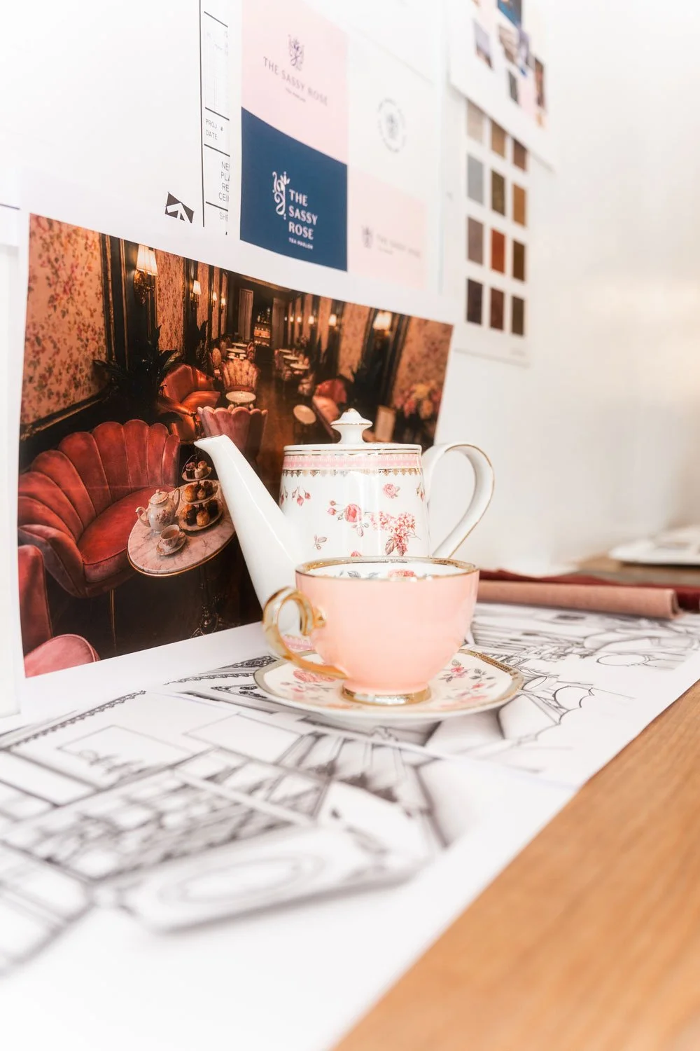 A pink and white floral teapot and a matching teacup with a saucer, placed on a table with sketches and design images in the background, including a mockup of the interior design
