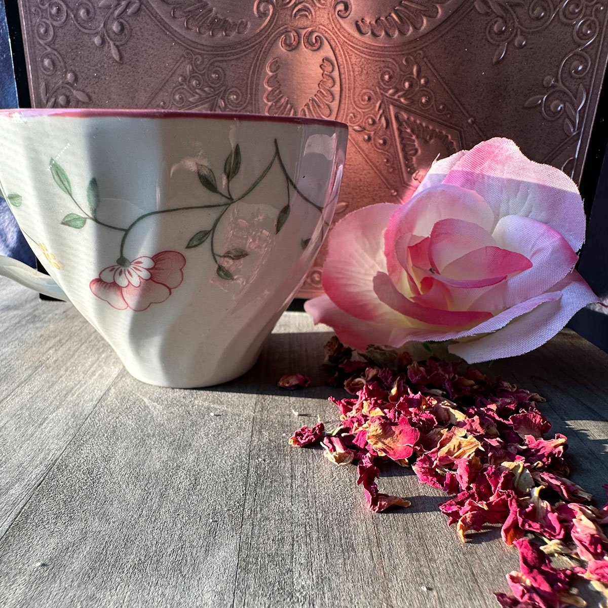 A white porcelain teacup with pink and green floral patterns, next to a pink flower and dried flower petals