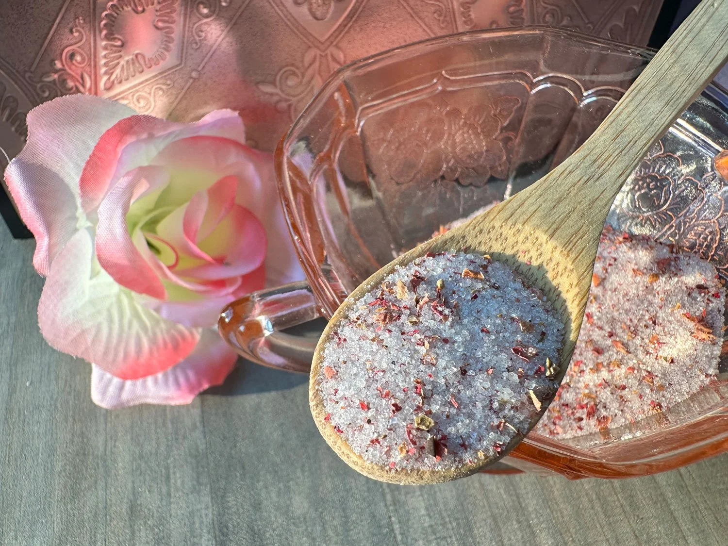 A pink and white flower, a glass bowl, and a wooden spoon filled with pink Himalayan salt