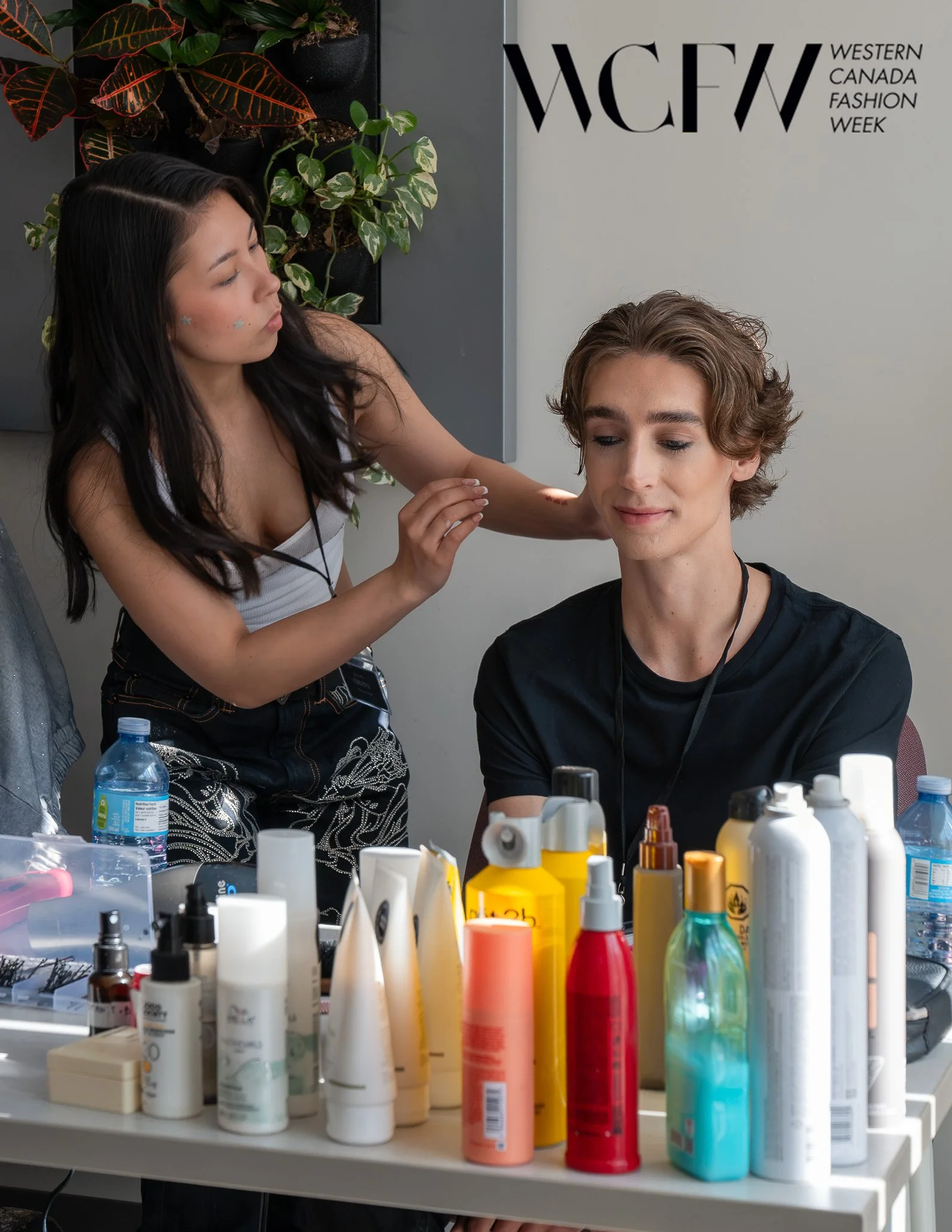A woman getting her makeup done by a makeup artist at Western Canada Fashion Week (WCFW). The makeup artist is working on the woman’s face, which is smiling slightly, and there are various makeup products and bottles on the table in front of them.