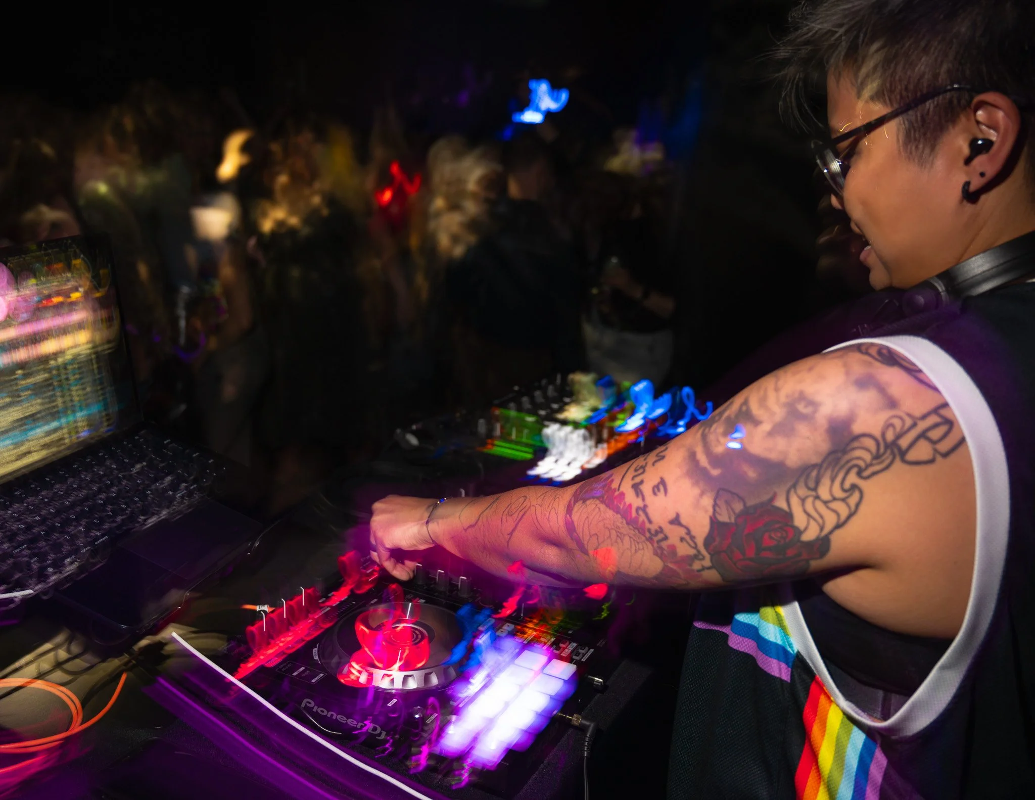 A DJ with tattoos on their arm, wearing glasses, mixing music on a Pioneer DJ controller at a party or club with colorful lights and a crowd.