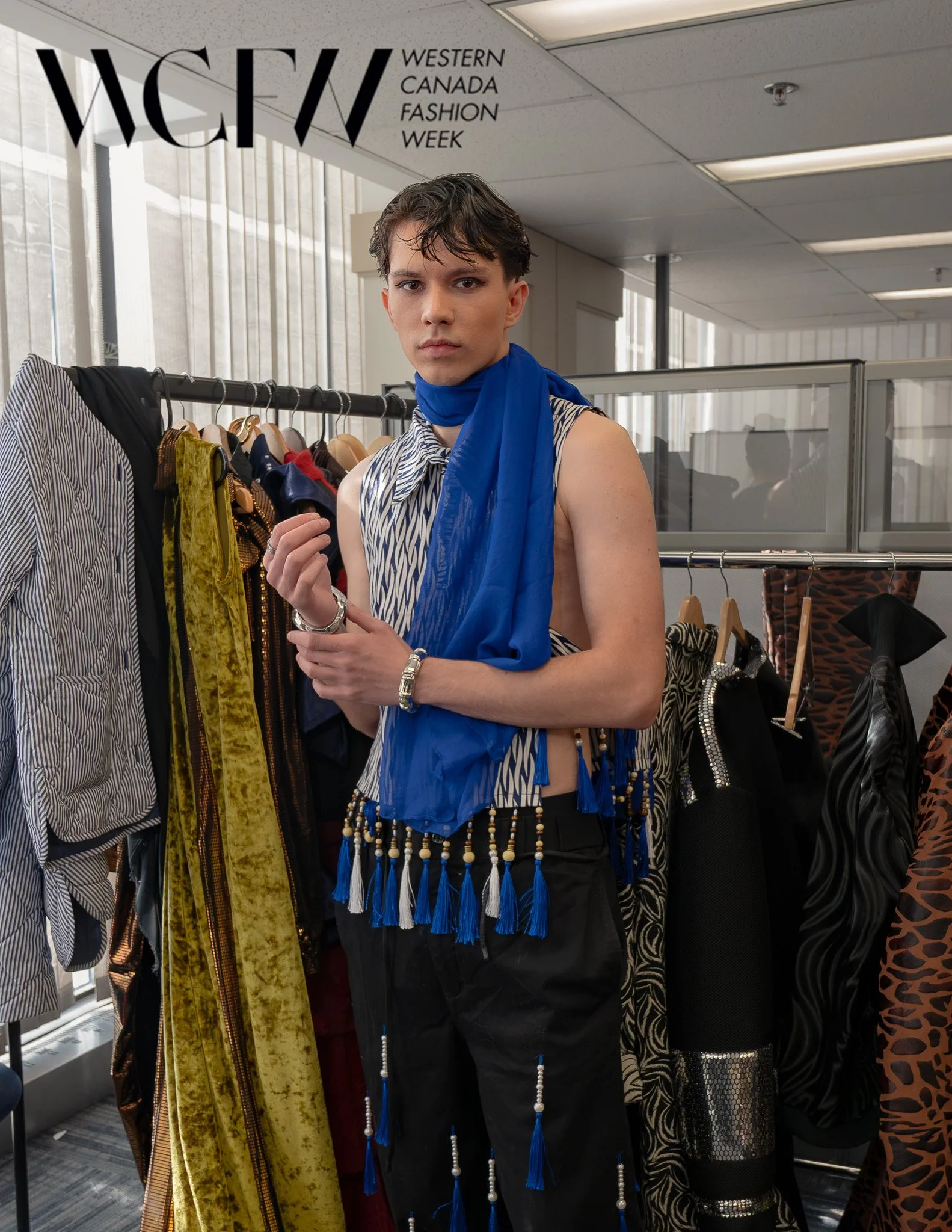 A young person with short dark hair and makeup, wearing a blue tassel necklace and earrings, standing in front of a clothing rack at Western Canada Fashion Week. The person is holding a piece of clothing or accessory and looking directly at the camer