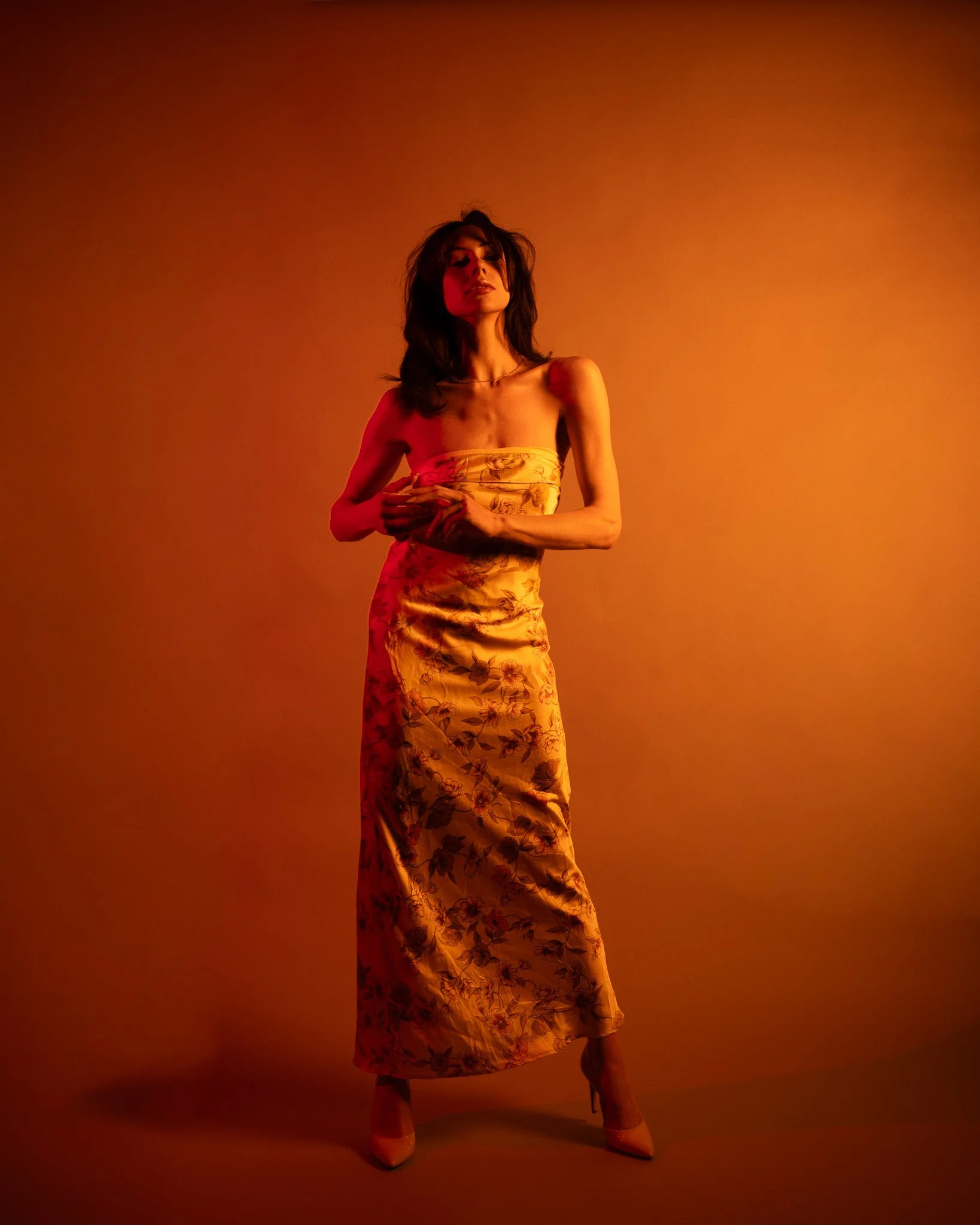 A woman in a floral strapless dress and beige heels stands against an orange background with lighting casting red hues on her face and shoulder.