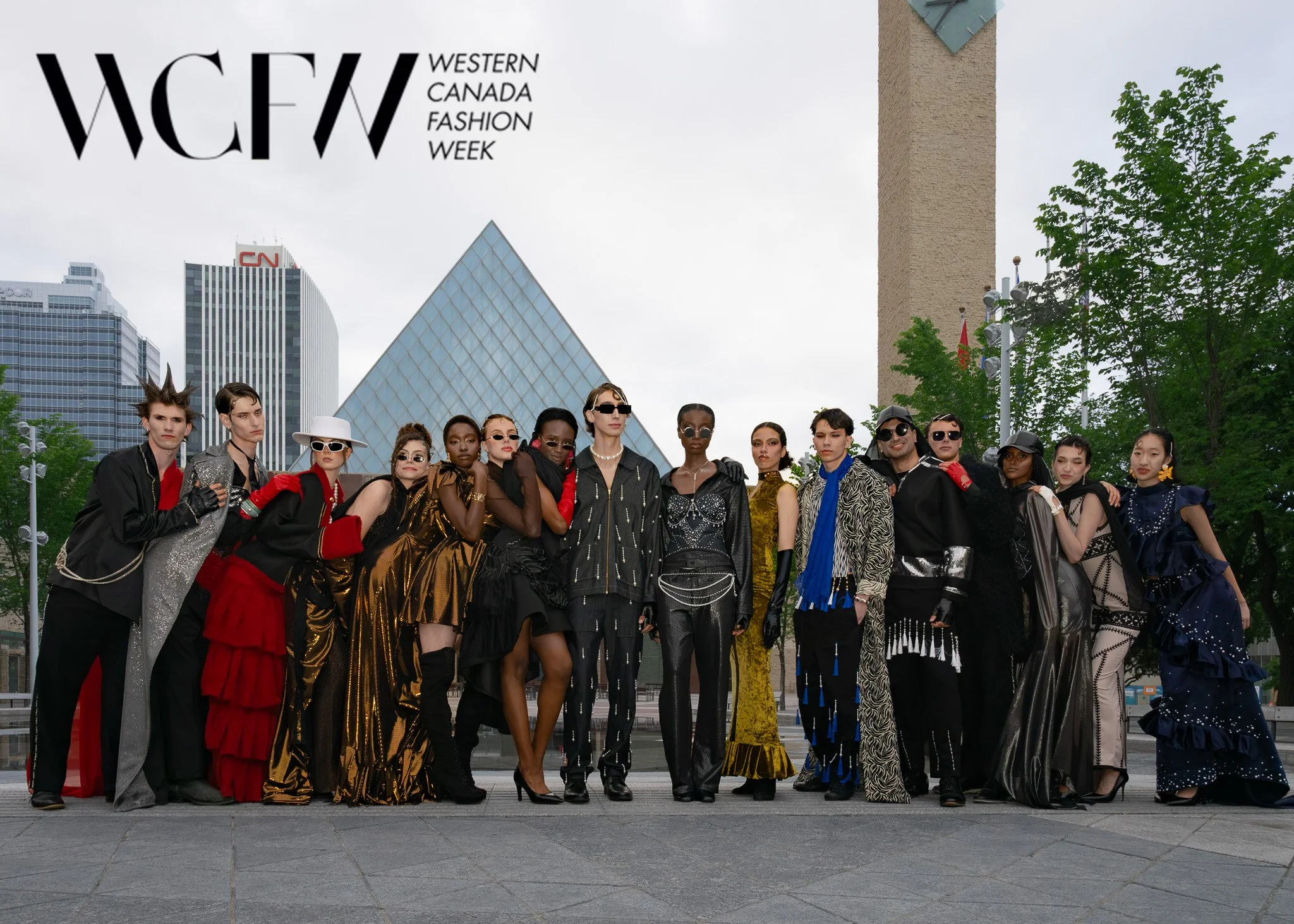 A group of models wearing fashionable clothing poses outdoors in front of city skyscrapers during Western Canada Fashion Week. The models are diverse in appearance, standing together on a paved area with trees and modern buildings in the background.