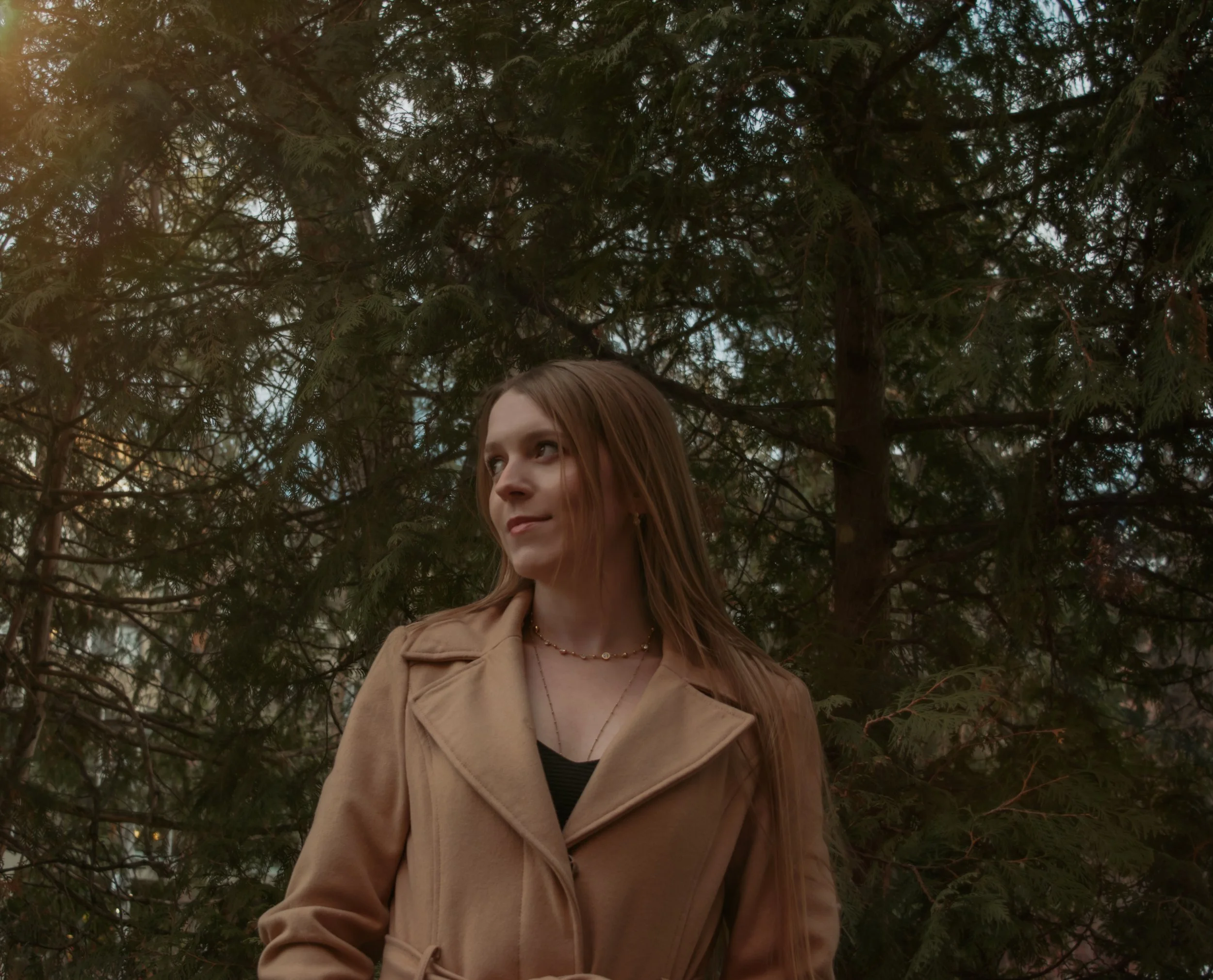 A young woman with long red hair wearing a beige coat and layered necklaces stands outdoors in front of pine trees.