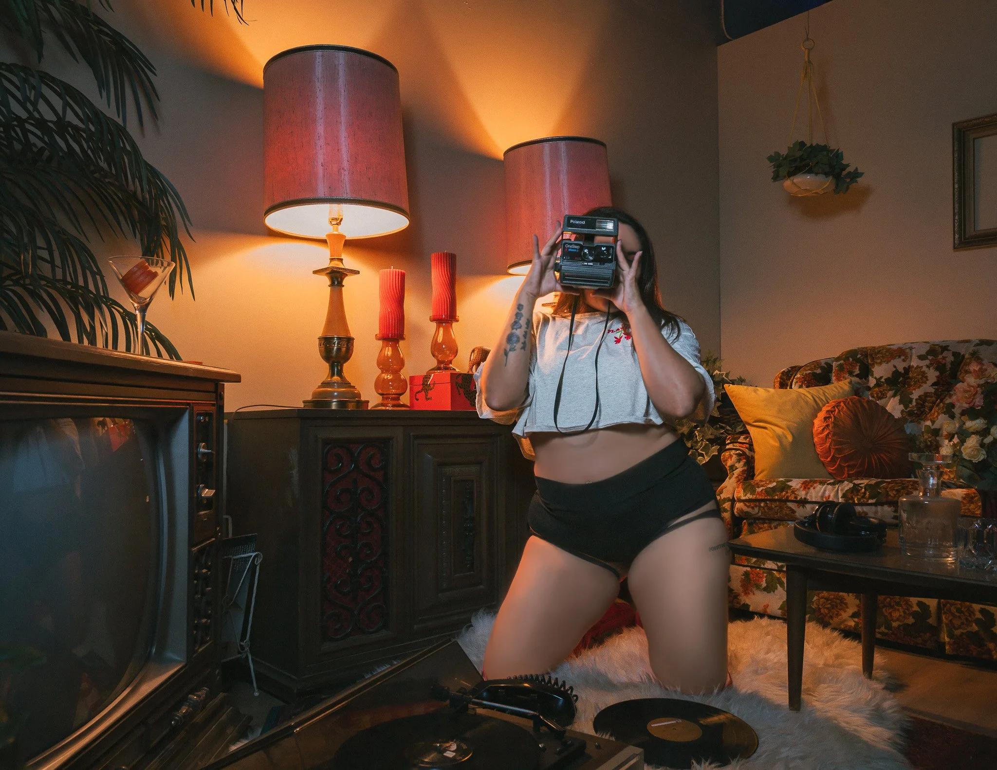 A woman in black shorts and a cropped t-shirt taking a photo with a vintage camera in a cozy, warmly lit living room with floral and plain cushions, a potted plant hanging from the ceiling, and a lamp on a sideboard.