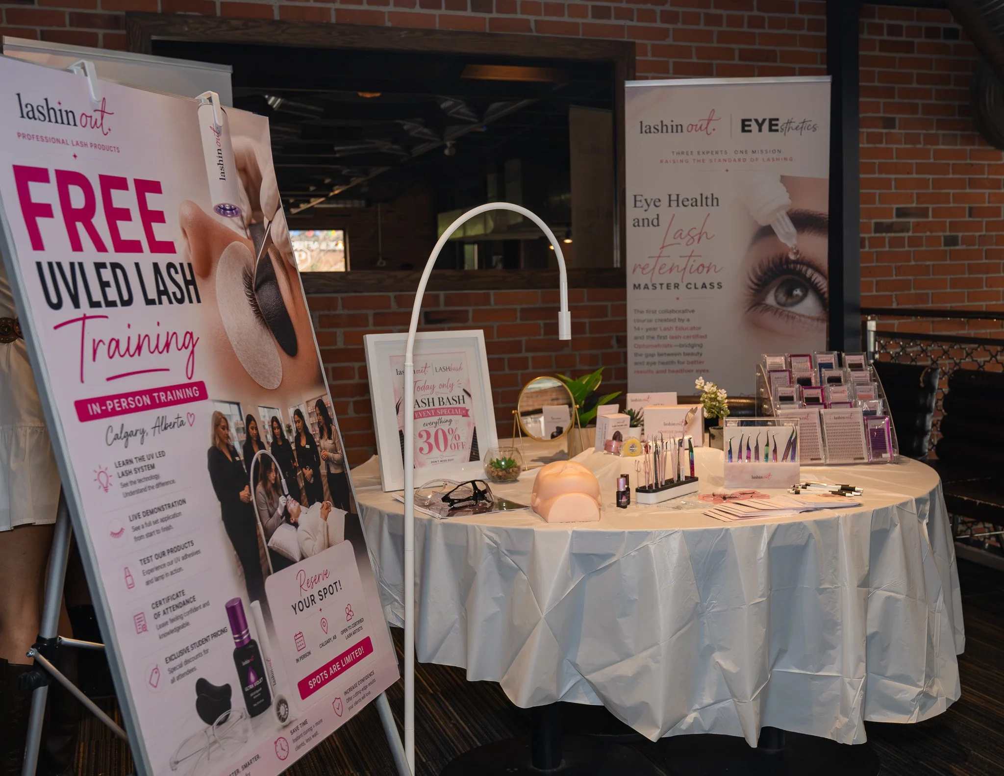 Display at a beauty event showing promotional posters and products related to eyelash extensions and eye health, in a venue with brick walls. The posters promote free eyelash training, an event special with 30% off, and a master class on lash retenti