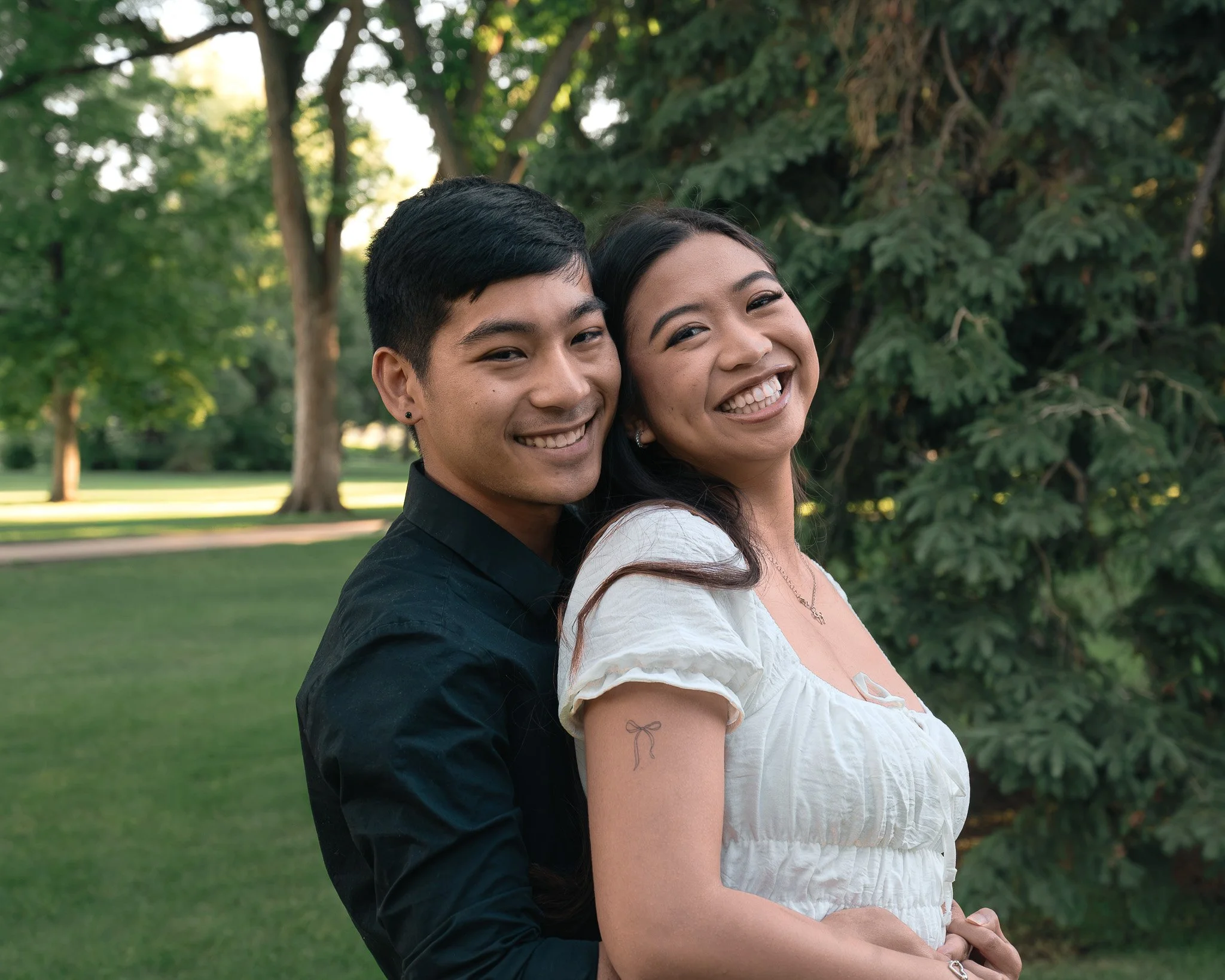 A young man and woman are standing close together outdoors in a park, smiling at the camera. The man has short black hair, wearing a black shirt, and the woman has long dark hair, wearing a white top.
