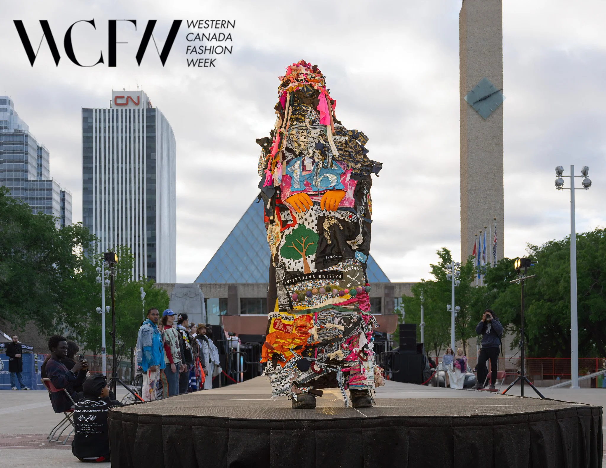 A person dressed in a costume made of colorful fabric pieces stands on a stage during Western Canada Fashion Week. The background shows tall buildings and a cloudy sky.