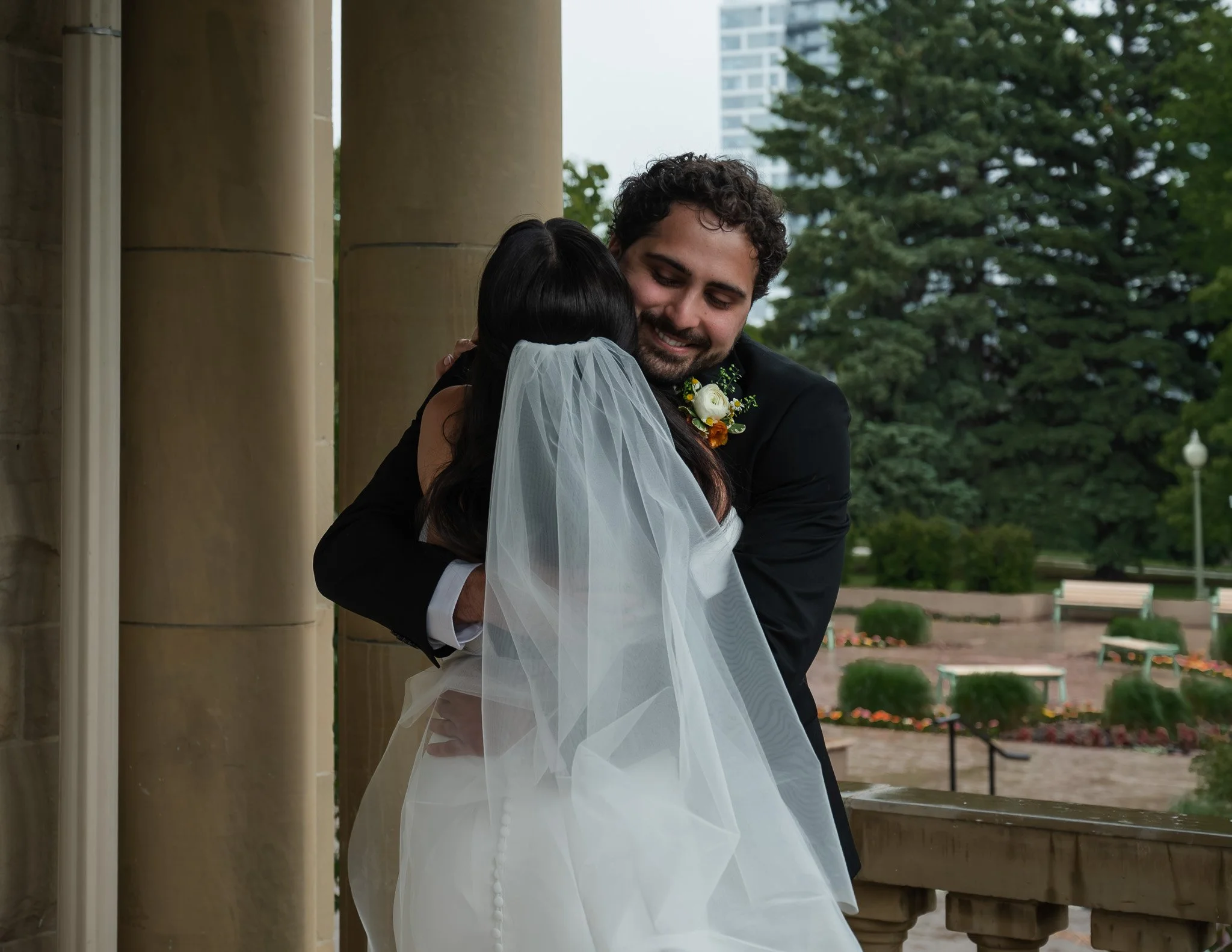 A man and woman, both dressed in formal attire, are sharing a hug outdoors during a wedding. The woman is wearing a white wedding dress and veil, and the man is wearing a black suit with a boutonniere. There are trees and buildings in the background.