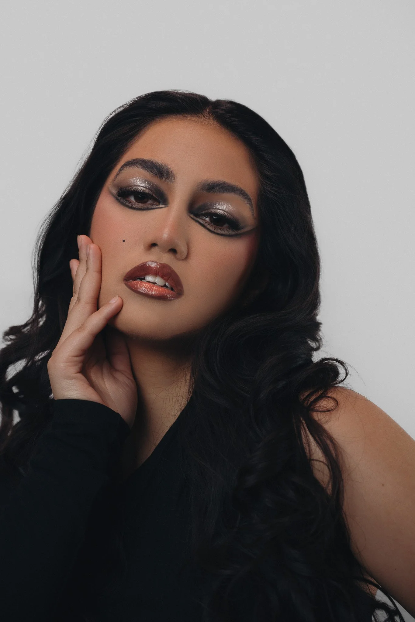 Close-up of a woman with long black hair, smoky eye makeup, glossy lips, and a beauty mark, holding her face with one hand against a plain background.
