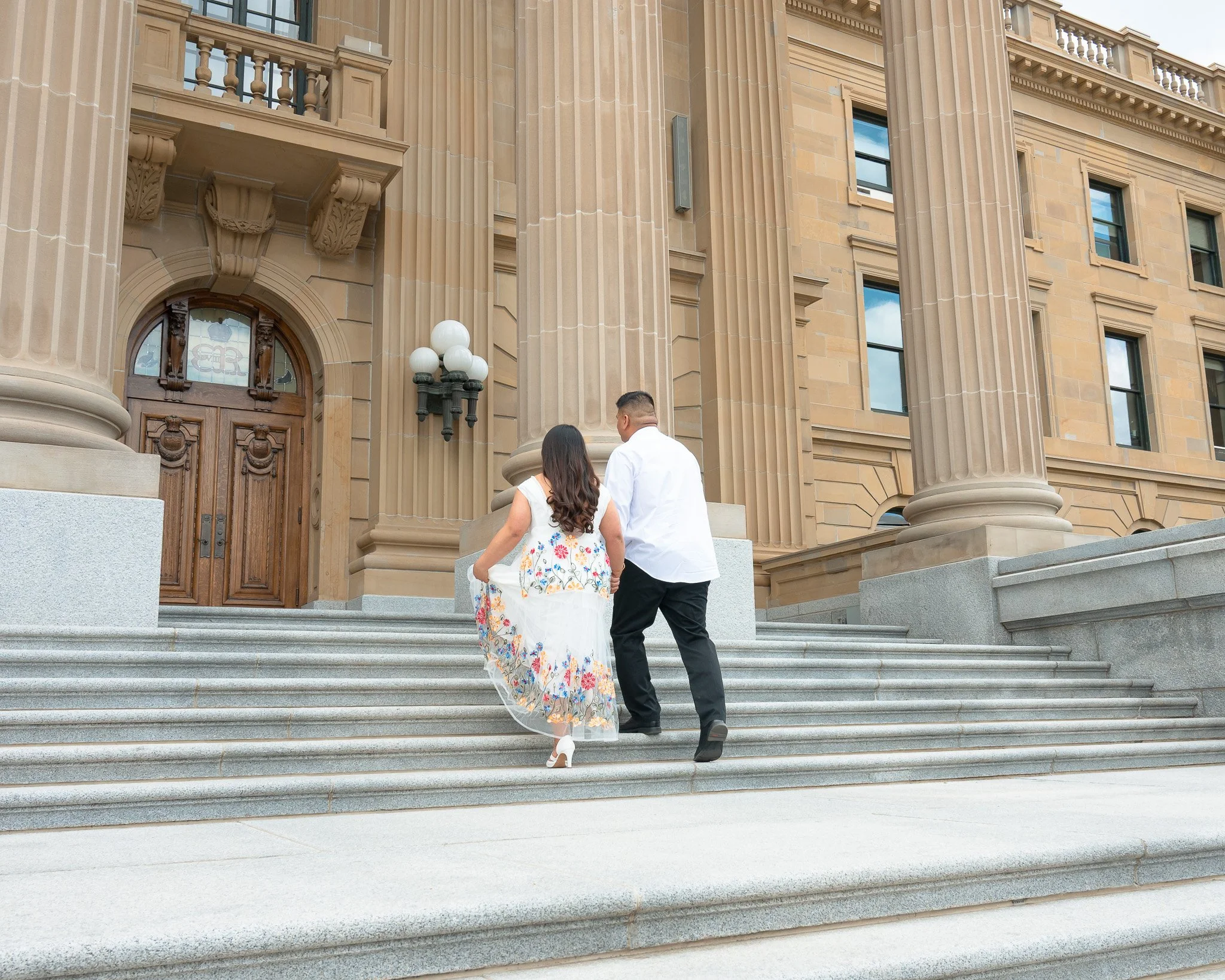 A couple dressed in wedding attire walking up the steps of a grand, historic building with large columns and ornate architectural details.