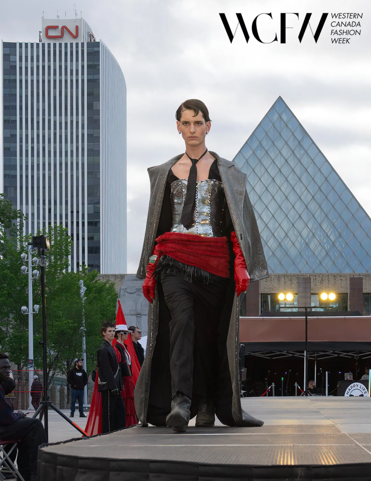 A runway model with short dark hair and makeup, wearing a metallic top, red gloves, a black tie, and a long sparkly coat, walking on an outdoor stage at Western Canada Fashion Week. There are tall buildings in the background, including a CN Tower, an