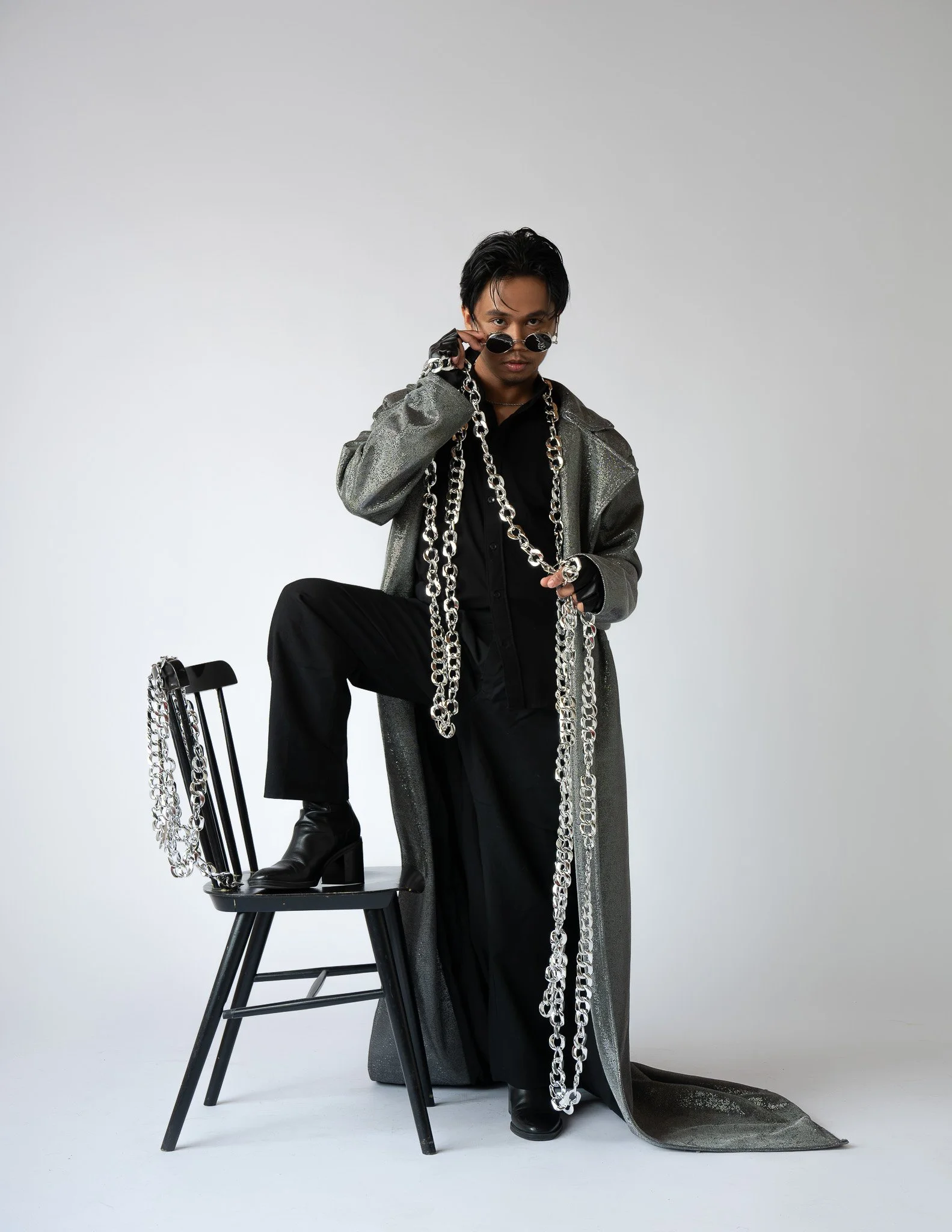 A person dressed in black and metallic gray clothing, wearing sunglasses, with multiple large silver chains, posing with one foot on a chair and the other on the floor, against a plain light gray background.