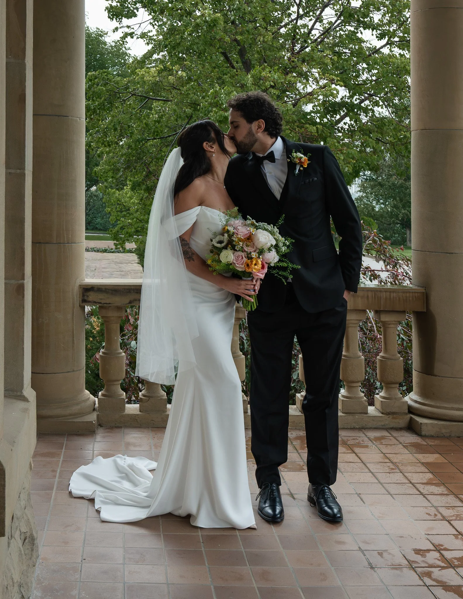 A bride and groom kissing on a balcony with columns and green trees in the background. The bride is wearing a white wedding dress and veil, holding a bouquet of flowers. The groom is dressed in a black tuxedo with a boutonniere.