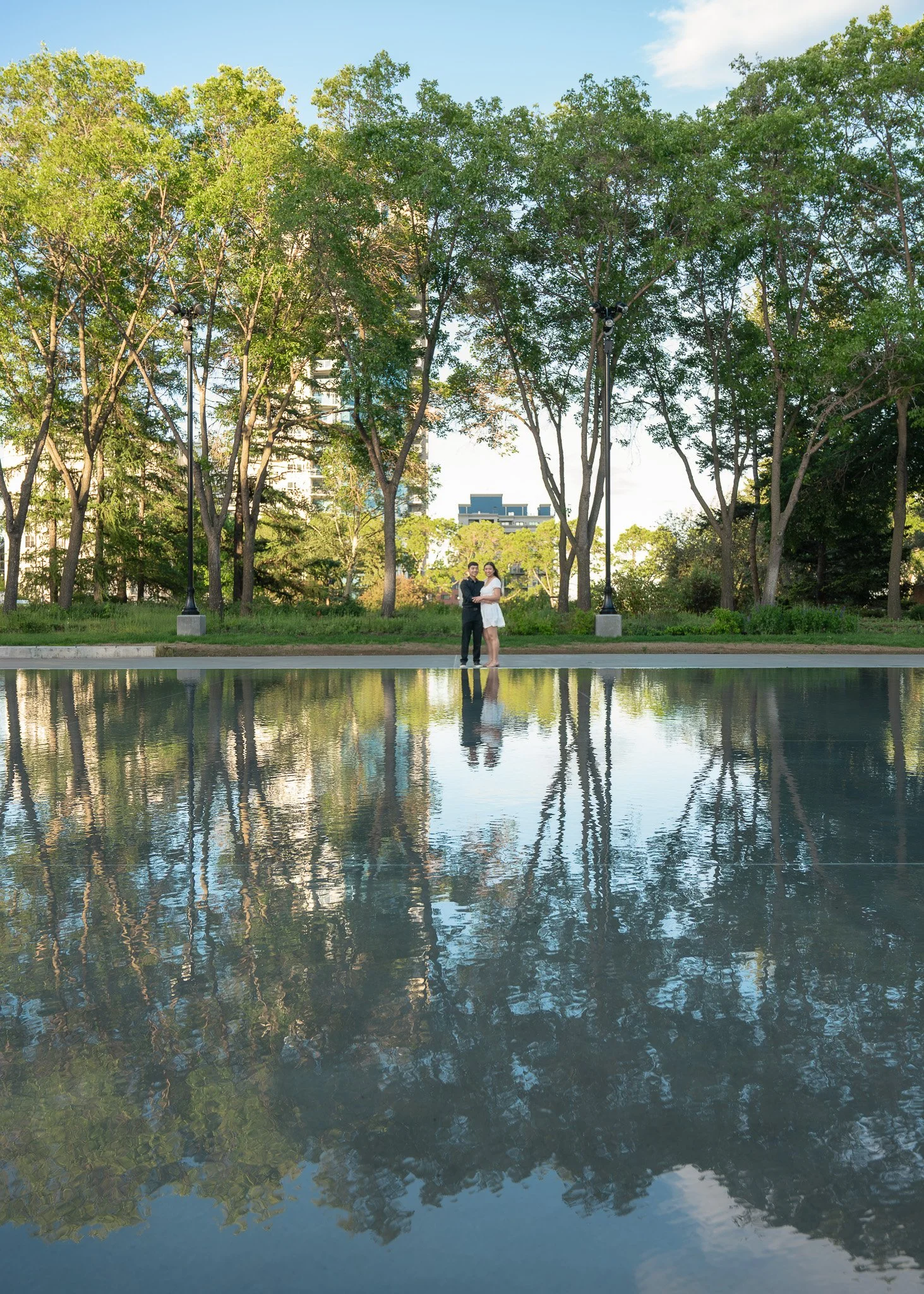 A man and woman dressed in formal attire standing on a sidewalk next to a body of water with trees and city buildings behind them, reflected in the water.