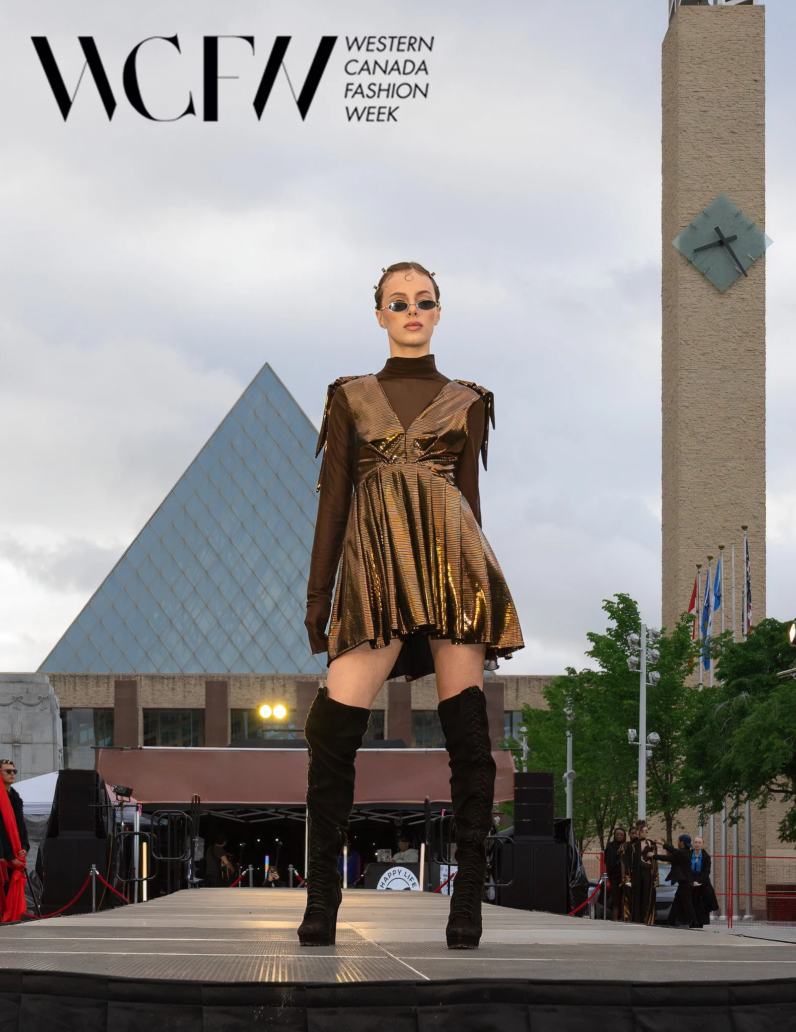 Model standing on stage at Western Canada Fashion Week, wearing a shiny gold and black dress with thigh-high black boots, sunglasses, and slicked-back hair, outdoors with a cityscape background.