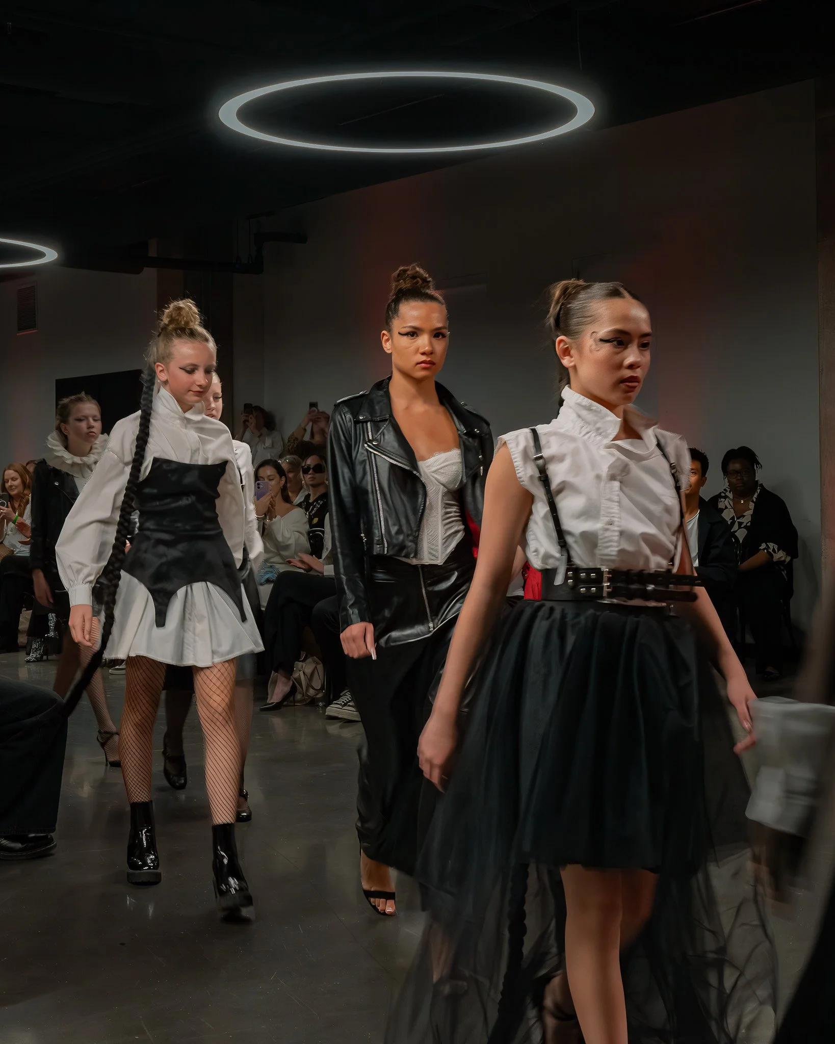 Models walking down the runway during a fashion show, wearing high fashion outfits with dark ambient lighting and circular ceiling lights.